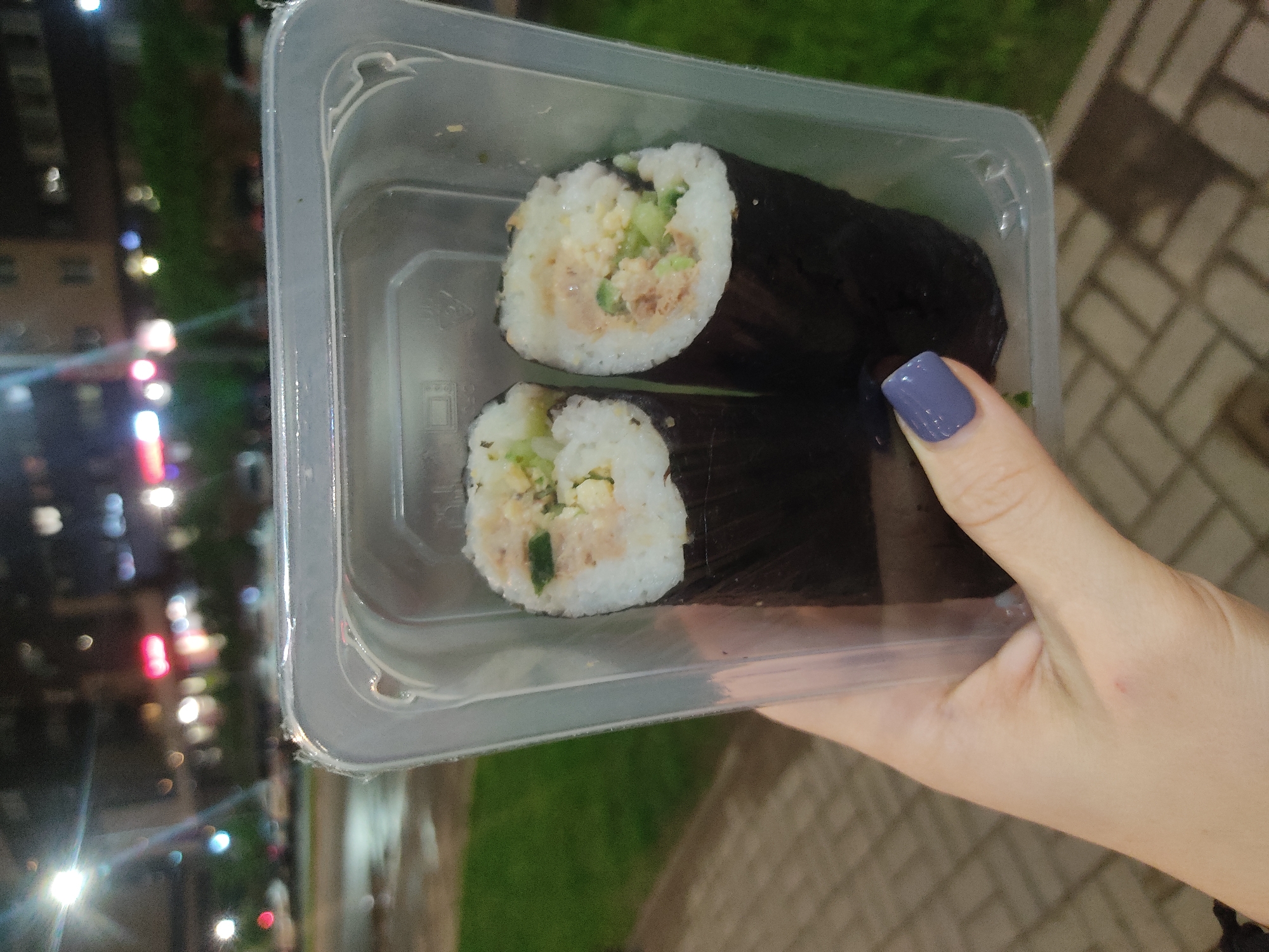 A hand holds a clear, rectangular plastic container containing two pieces of sushi rolls. The sushi appears to be made with white rice, nori seaweed, and fillings that include tuna, cucumber, and egg. The fingernail of the hand holding the container is painted a bluish-grey color. The background shows an urban environment with blurred lights from buildings and some greenery, suggesting an outdoor location. The scene appears to be set in the evening or at night. A brick path or pavement is also visible in the background.