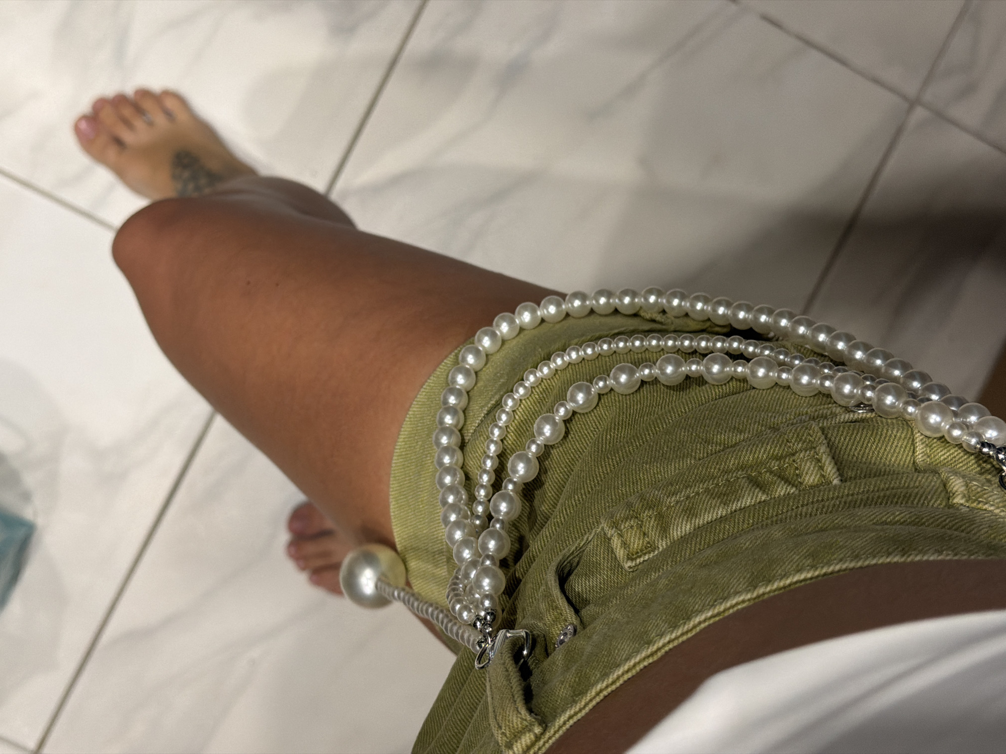 A close-up, overhead shot shows a person's legs and a string of pearls draped over a pair of olive-green shorts. The person's right foot is visible, and they have a tattoo on their ankle. The background consists of white tiled flooring. The image does not contain any identifiable landmarks or elements to suggest a specific location in Irkutsk, Russia.