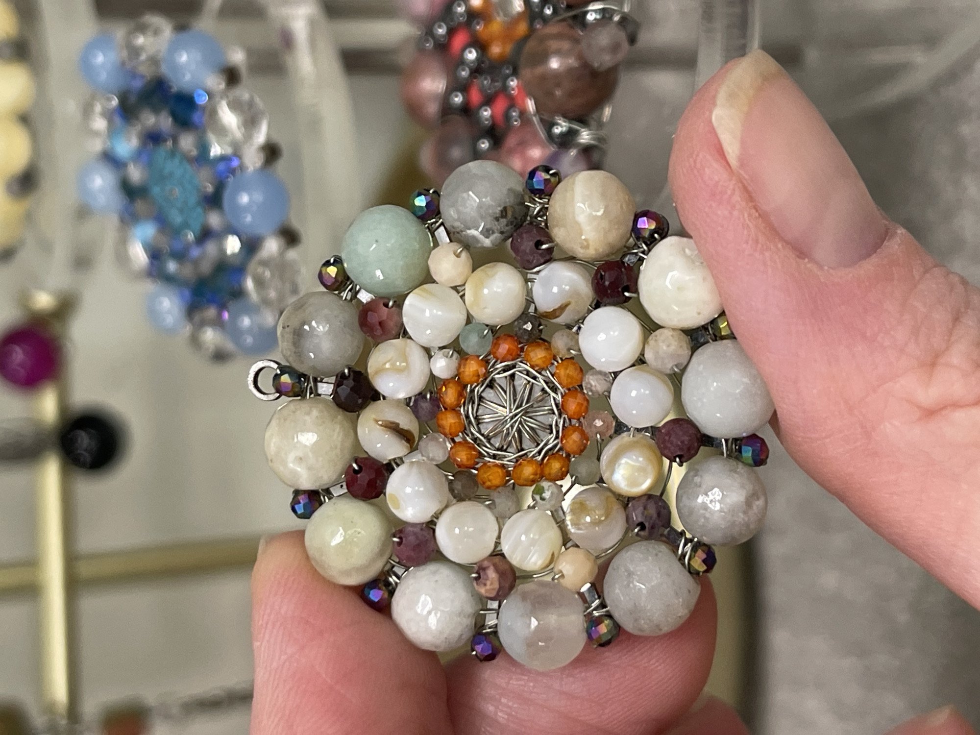 A close-up shows a hand holding a circular, beaded brooch. The brooch has a variety of round beads in different sizes and colors, arranged in a floral-like pattern. The colors include white, beige, gray, orange, and purple. The center features a metallic, wired structure. Iridescent, faceted beads are spaced around the larger beads. The background is blurry and appears to show other beaded pieces. The hand is holding the brooch with the thumb, which is in focus. The image location is not discernible in the provided scene.