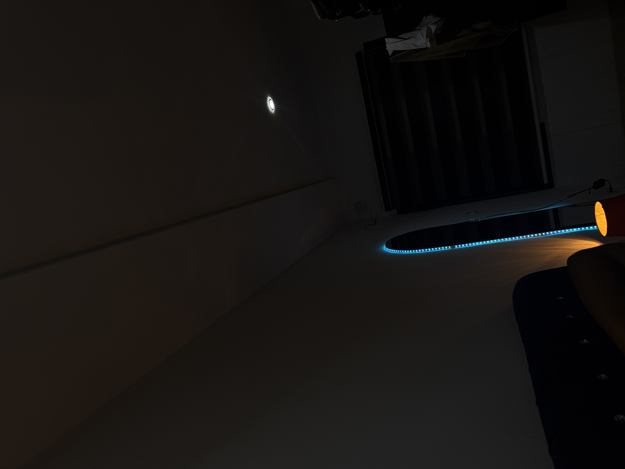 The image depicts a dimly lit interior scene. A dark wall is visible with a recessed light fixture emitting a soft glow. In the foreground, a black object with reflective embellishments is partially visible. A circular light fixture with blue LEDs illuminates a portion of the ceiling. In the background, a window or doorway with dark vertical blinds is present. The scene could be located in Abuja, Nigeria, but there is no information in the photo to confirm that.