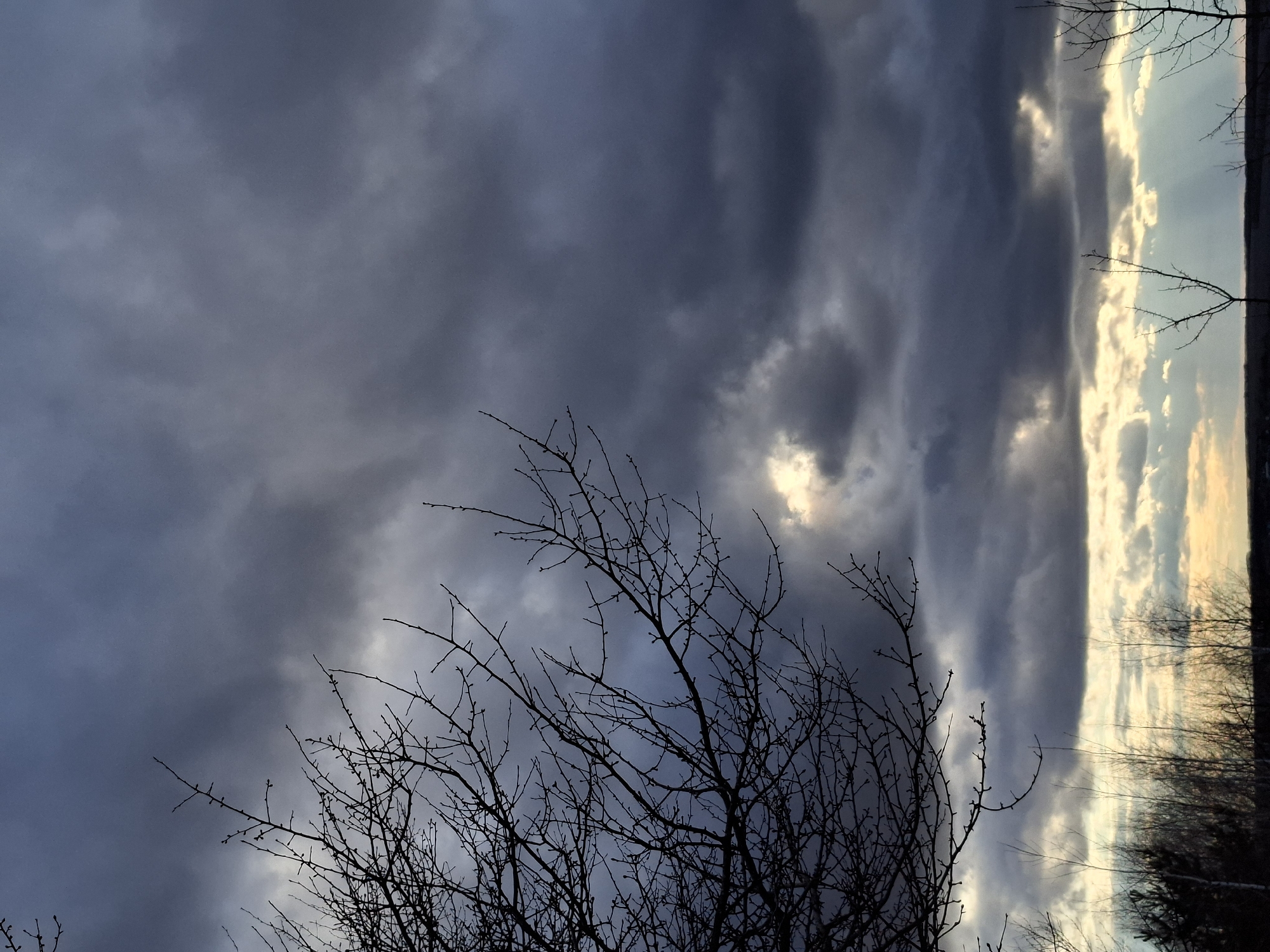 The image shows a view of the sky from Khlevnoye, Russia. The sky is filled with dark, heavy clouds, with a small patch of light visible between them. The silhouette of a bare tree is visible in the foreground. The scene is mostly dark, with muted colors.