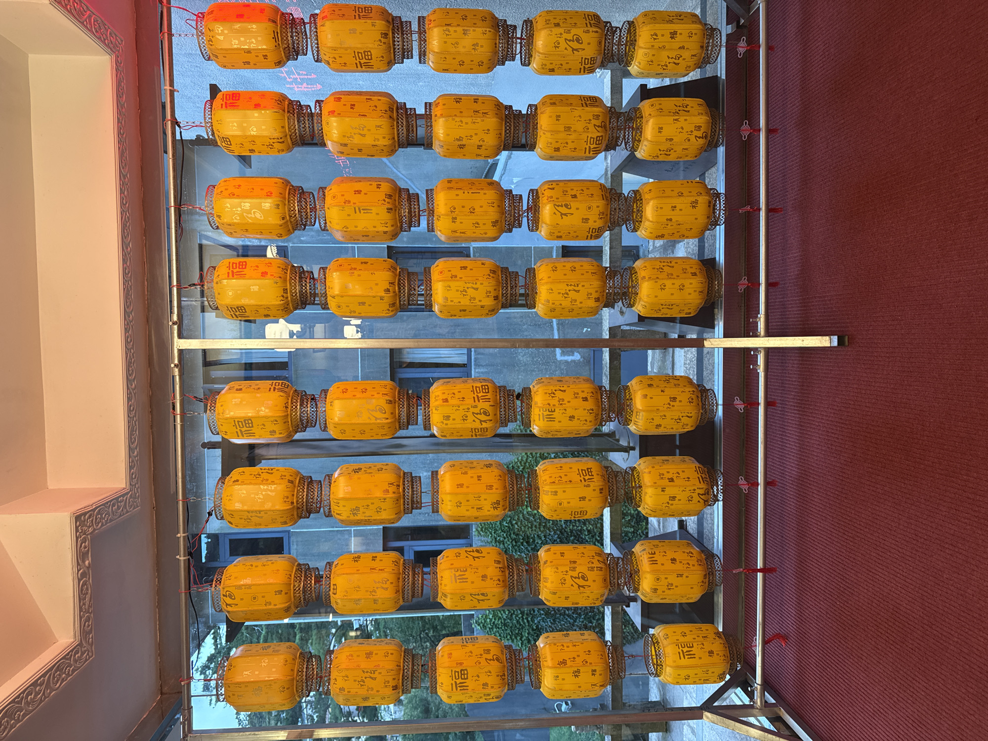 A row of yellow lanterns, adorned with Chinese characters, are suspended in front of a building. The lanterns are arranged in vertical columns, with multiple lanterns stacked on top of each other. The building behind them appears to be made of concrete and glass, with windows visible. The scene is illuminated by artificial light, suggesting it is nighttime or an indoor setting. The lanterns are decorative and likely part of a festive display. The ground in the foreground is covered with a red carpet. The overall impression is one of cultural celebration and decorative ambiance. The location is the city of Huangshan District, China.