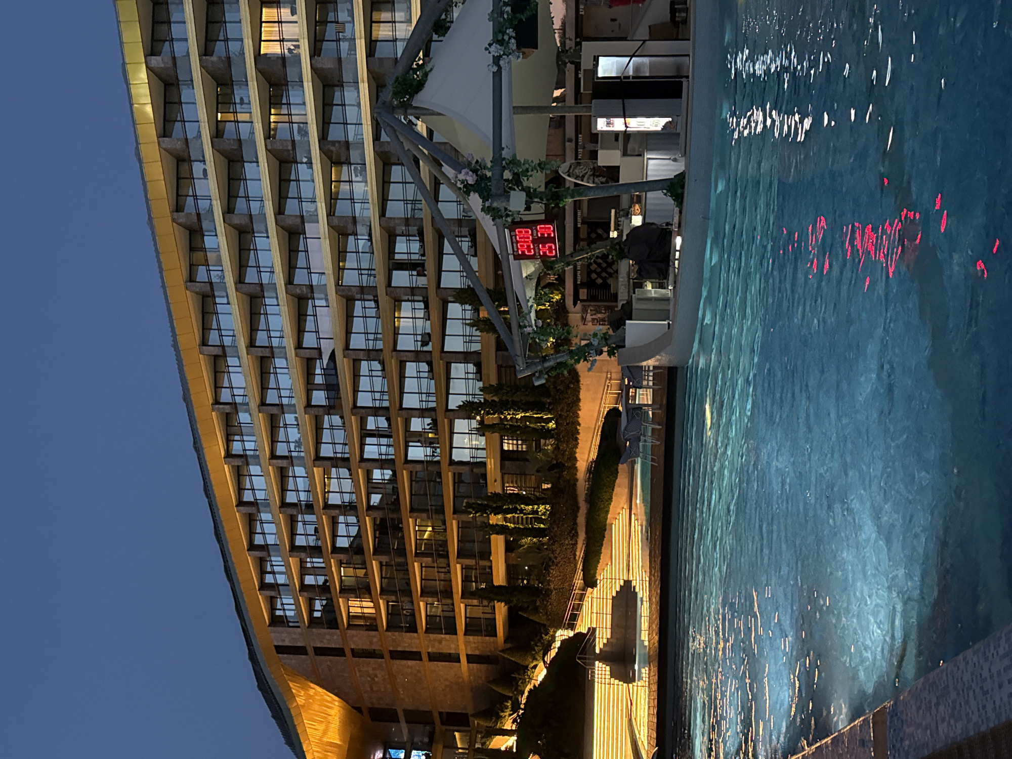 This image shows a modern hotel building at dusk, situated next to a swimming pool. The hotel, with its multi-story balconies and illuminated windows, appears to be of contemporary design. The swimming pool is illuminated, reflecting the lights from the hotel and some red lights from a structure at the pool's edge. A small bar or service area is visible near the pool, featuring a digital display showing "29.08" and "25 30". The surrounding environment includes landscaped terraces with stairs leading up to the hotel and some trees. The sky is a deep blue, indicating twilight. The overall scene suggests a resort or hotel complex. The location is identified as Симеизский поселковый совет, Ukraine.
