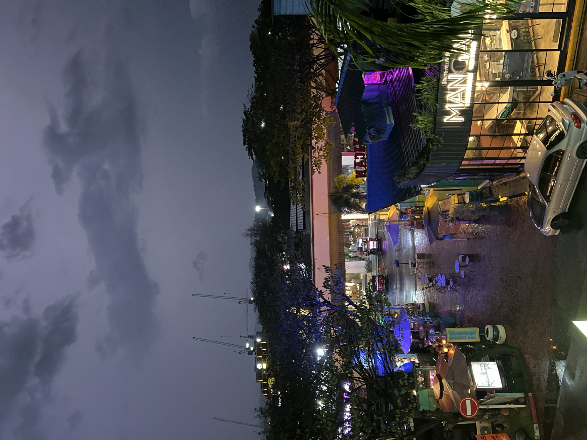 Here is a description of the image:
The image shows a city street at night, possibly after a rainfall, given the wet pavement. The scene is lit by streetlights and neon signs from businesses. The dominant structure is a restaurant or bar with a large, illuminated sign that reads "MAN". It has a curved glass facade with lights on inside. Cars are parked nearby, and outdoor seating is visible, indicating a public space for dining or socializing. The sky is dark and cloudy, and the construction cranes loom in the background, suggesting ongoing urban development in the area. In the foreground, there is a tree and some signage, including what appears to be a no-entry sign. People are present at the cafe's tables and on a motorcycle with a trailer. The location is in the city of Unknown, Unknown.