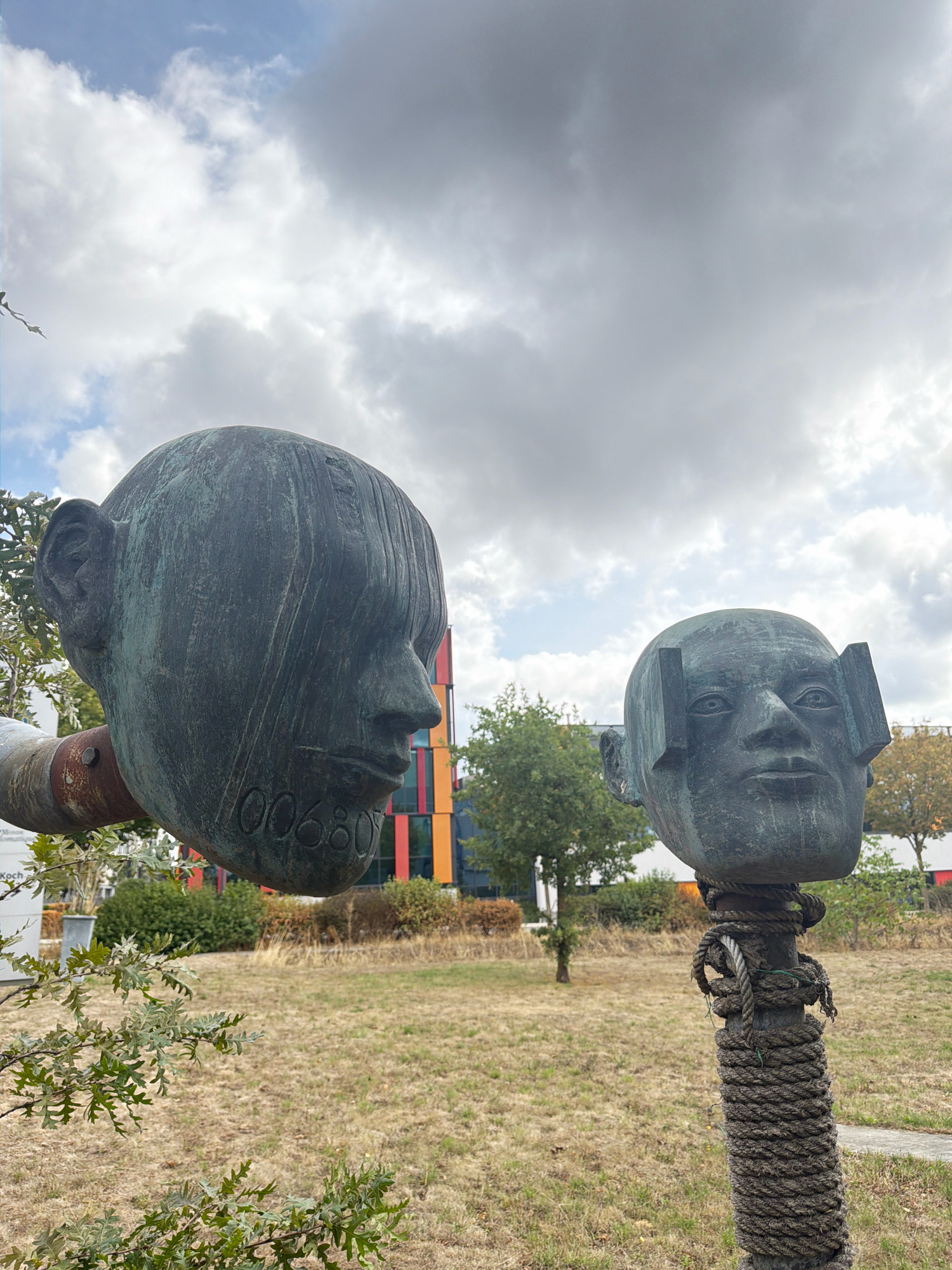 The image shows two bronze head sculptures outdoors in Esch-sur-Alzette, Luxembourg. The sculpture on the left is larger, with a detailed ear and a numerical inscription of "00680" on its cheek. It is attached to a rusty pipe that extends from the left edge of the frame. The sculpture on the right is smaller and features two rectangular protrusions on either side of its forehead, obscuring the eyes. This sculpture is mounted on a pole wrapped with thick rope.
In the background, a modern building with colorful vertical panels in red, orange, yellow, and blue is visible. The environment consists of dry, brown grass, scattered green bushes, and several trees. The sky is overcast with large, grey clouds. The overall scene suggests an outdoor art installation in a park or public space.