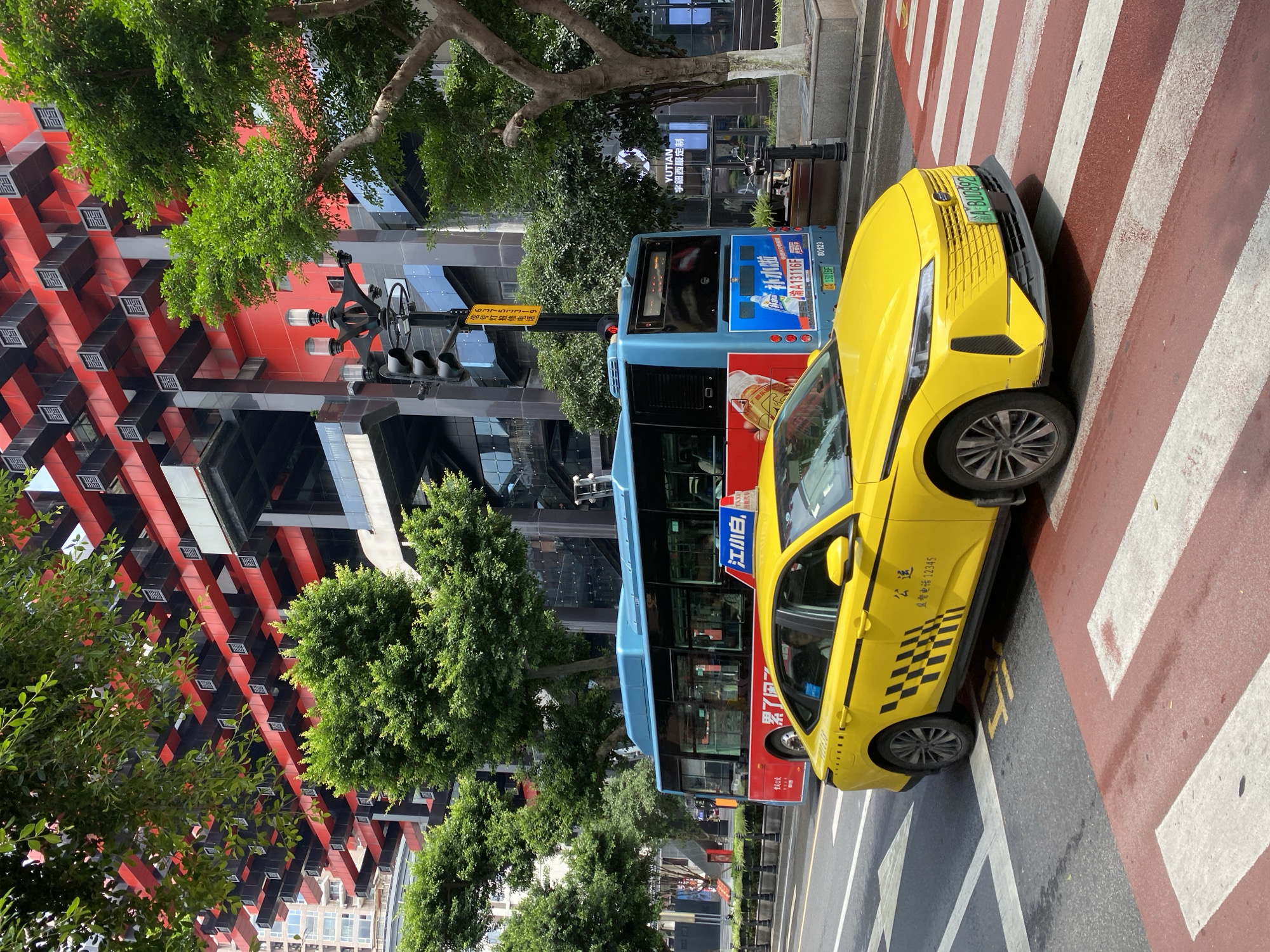 This media file captures a street scene in Yuzhong District, China, likely during the daytime given the bright sunlight. A bright yellow taxi is prominently featured in the foreground, positioned on a pedestrian crossing with red and white stripes. The taxi has "公运 监督电话12345" (Public Transport Supervision Phone 12345) written on its side, along with a checkered pattern near the rear wheel and the license plate "A BU0892".
Behind the taxi, a blue public bus is visible. It displays advertisements, including one with Chinese text and an image of a drink. The bus's license plate reads "A13116F".
The background reveals a modern urban environment. A striking building with a red and black geometric facade stands to the left, interspersed with trees and greenery. Another building with reflective glass windows can be seen further back. A traffic light signal with multiple lamps is also present. The scene suggests a busy city street with both public and private transportation. The overall impression is one of a typical urban day in China.