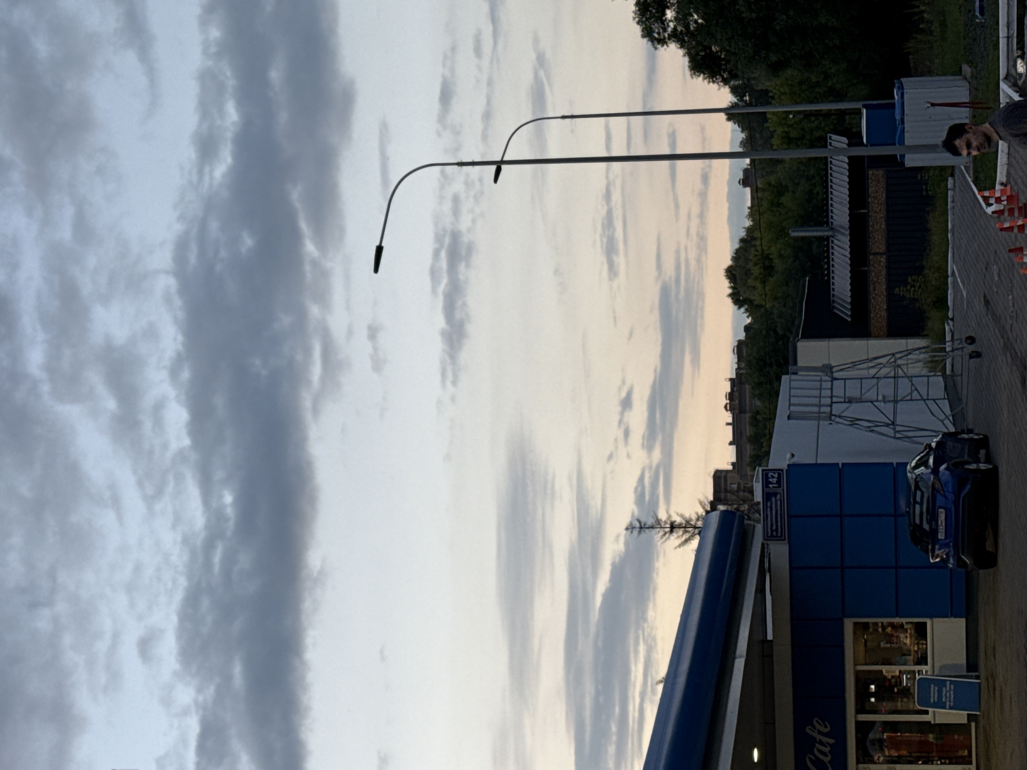 The image shows an outdoor scene at dusk, likely at a gas station with an attached cafe. In the foreground, a dark blue car is parked near the building. The building has blue and white elements, with a sign that says "Cafe". To the right of the building, there is a man standing and looking towards the camera. He is wearing a dark t-shirt. In the background, there are trees, streetlights, and buildings. The sky is cloudy. There is a street sign visible with "142" on it. The overall impression is a common urban or suburban setting.