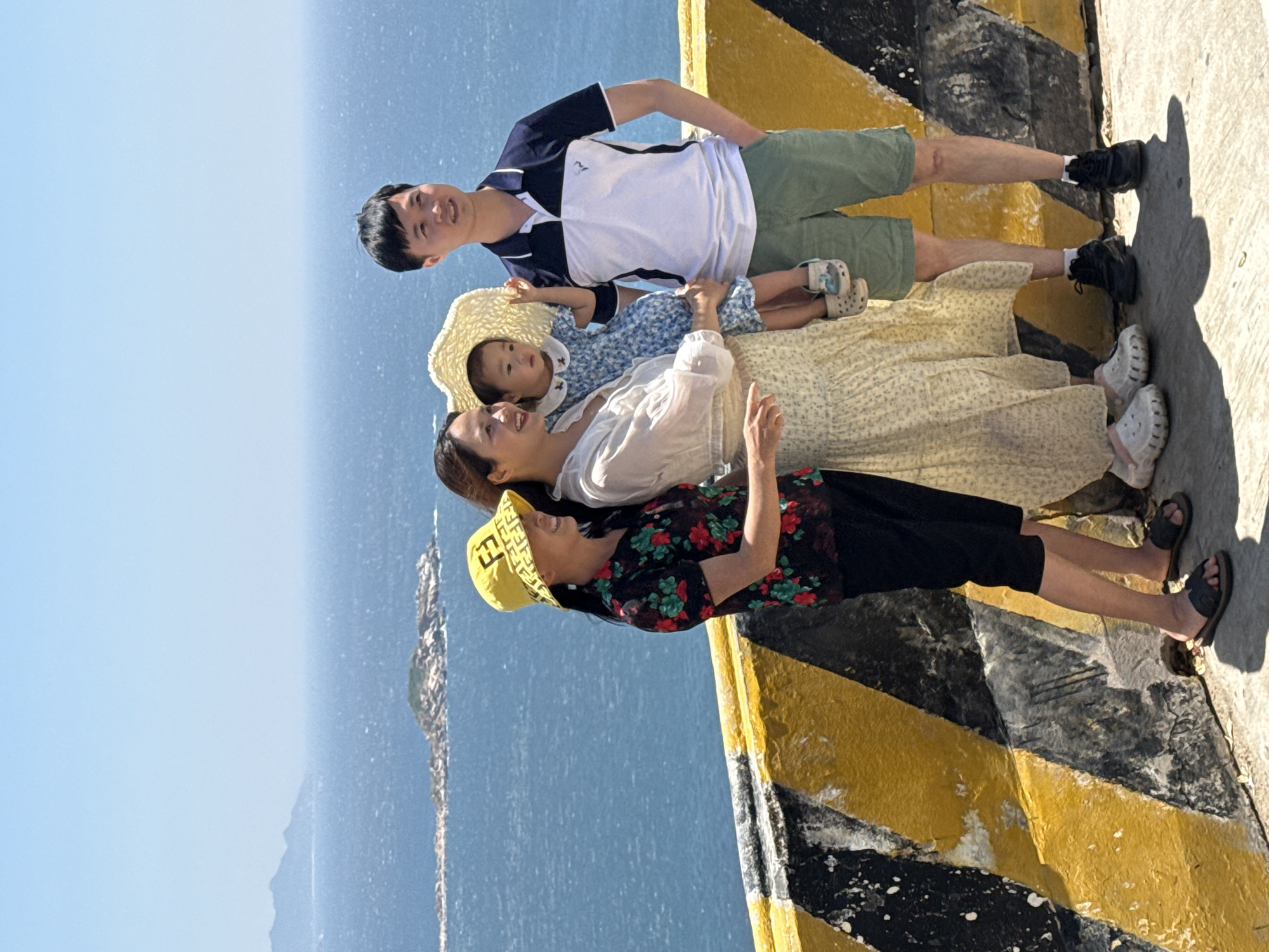 Four individuals, comprising three adults and one young child, are posing for a photograph outdoors at a scenic overlook in Vinh Hai, Vietnam. They stand in front of a concrete barrier painted with diagonal yellow and black stripes, overlooking a vast blue ocean under a clear sky.
The leftmost individual is an adult woman wearing a yellow bucket hat with a repeated pattern, a dark short-sleeved shirt with red and green floral print, and dark capri-style pants. She wears dark sandals and is smiling. To her right, a second adult woman holds a young child. This woman wears a light-colored, long-sleeved sheer blouse over a lighter-toned patterned dress and white platform clogs. She is also smiling and looking towards her left. The child, a toddler, wears a light blue floral dress with a white collar, a wide-brimmed straw hat, and white clogs. The child's right arm is slightly raised. To the right of the central woman and child is an adult man wearing a navy and white short-sleeved polo shirt, green shorts, and black sneakers with white socks. His left hand is in his pocket, and he looks towards the right.
In the background, the ocean stretches to the horizon. A long, rocky island or landmass is visible in the mid-distance on the water, with larger mountains or landforms faintly discernible on the far horizon to the left. Bright sunlight illuminates the scene, casting distinct shadows on the concrete ground in the foreground.