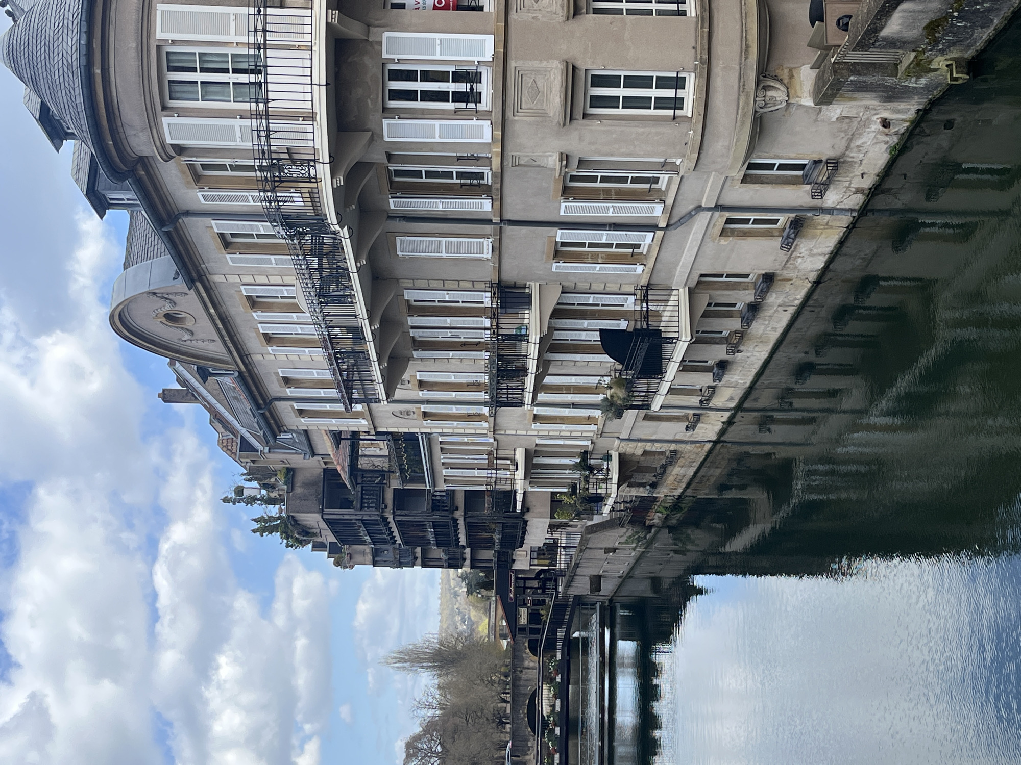 A building with multiple floors is reflected in the water of a canal or river. The building features numerous windows with white shutters, some of which are open and some are closed. Some windows have small wrought iron balconies. The building is located along the Quai des Éclusiers in Metz, France. A bridge spans the canal, and trees are visible along the embankment. The sky has scattered clouds.