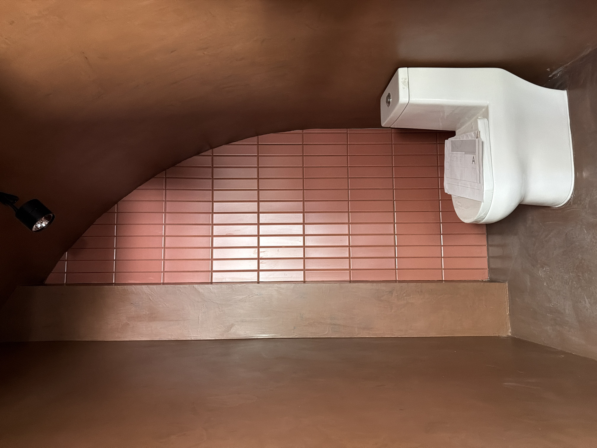 A medium-shot perspective looking up and into a space with an arched ceiling and earth-toned walls. A rectangular brick wall in a muted, reddish-brown tone is visible along the middle of the back wall. A white, irregularly-shaped object with a metallic dot on its left side and paperwork protruding from its underside is mounted on the right side of the back wall. A small, black, round spotlight is visible on the left wall, close to where the wall meets the arched ceiling. The scene suggests an interior space, possibly a modern architectural detail or design feature. The location is Puebla, Mexico.