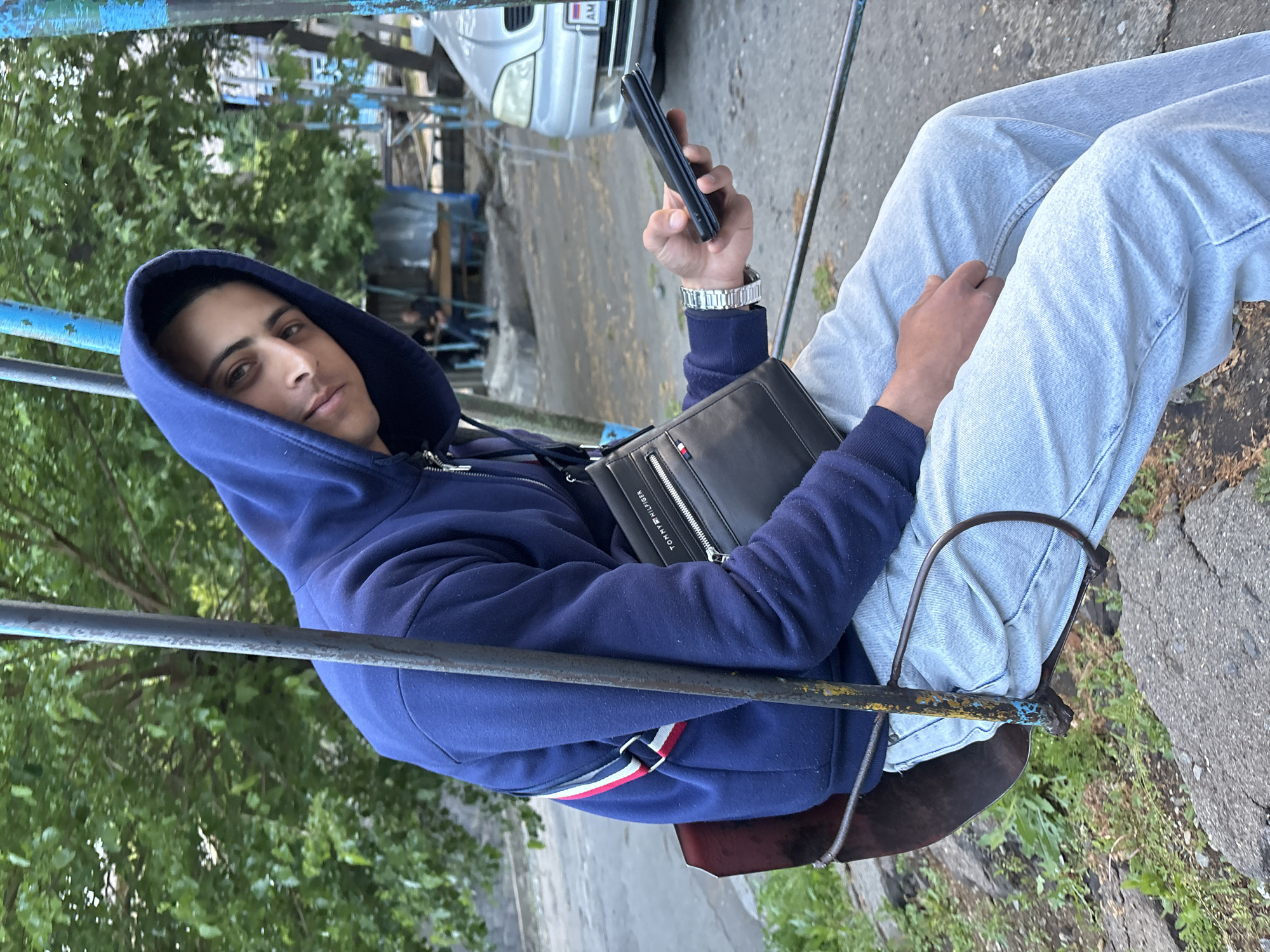 A person in a dark blue hooded sweatshirt and light blue jeans sits on a swing, holding a black smartphone in their right hand. They have a black Tommy Hilfiger bag slung across their body. The swing is blue with signs of wear. A silver car is parked in the background, with buildings and trees visible. The photograph appears to have been taken in Alaverdi, Armenia, based on the car's license plate.
