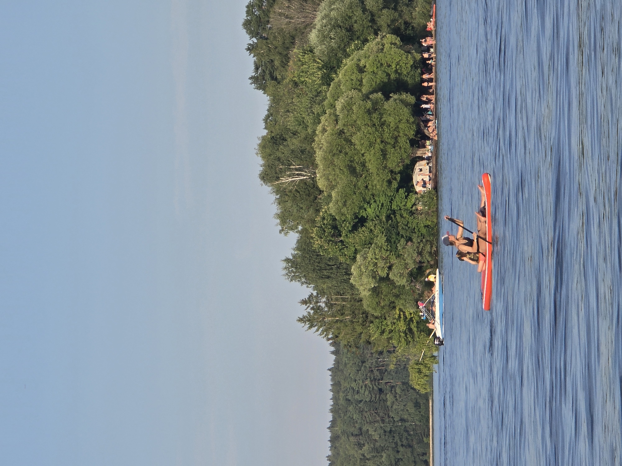 A wide-angle, medium shot captures a serene lake scene under a clear blue sky. In the foreground, a person on a bright orange paddleboard is paddling across the calm, blue water. The individual is wearing a dark swimsuit and a cap, and is actively engaged in rowing. In the mid-ground, further out on the lake, a white motorboat with several passengers is visible, moving away from the shore. The background is dominated by a lush, dense forest that lines the lake's edge, with a sandy beach area populated by a crowd of people enjoying the sunny weather. There are several tents and umbrellas set up on the beach, indicating a recreational area. The overall impression is a peaceful summer day at a popular outdoor destination, possibly in the vicinity of the Kuybyshev Hydroelectric Power Station settlement in Russia.