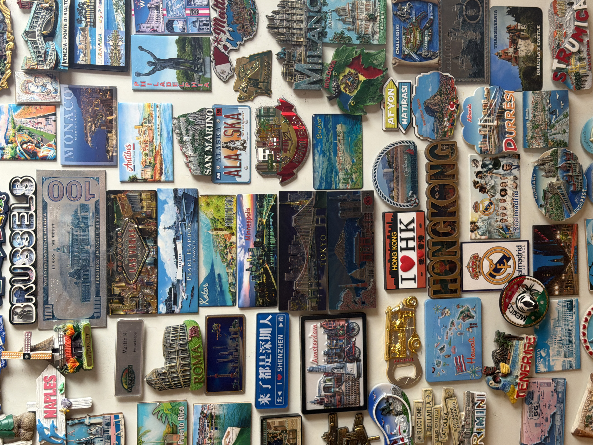 The image shows a collection of souvenir magnets attached to a white surface. The magnets represent various cities, countries, and regions around the world, including Venice, Monaco, Antibes, San Marino, Alaska, Brussels, Las Vegas, Puerto Rico, Rome, Kotor, Shenzhen, Amsterdam, Thailand, Hawaii, Tenerife, Mexico, Pearl Harbor, Philadelphia, Miami Beach, Tokyo, Hong Kong, Afyon, Durres, Transilvania, and Budapest.
There is no indication of Buhovo, Bulgaria, or any people or actions within the image. The scene is purely a static display of souvenir magnets.