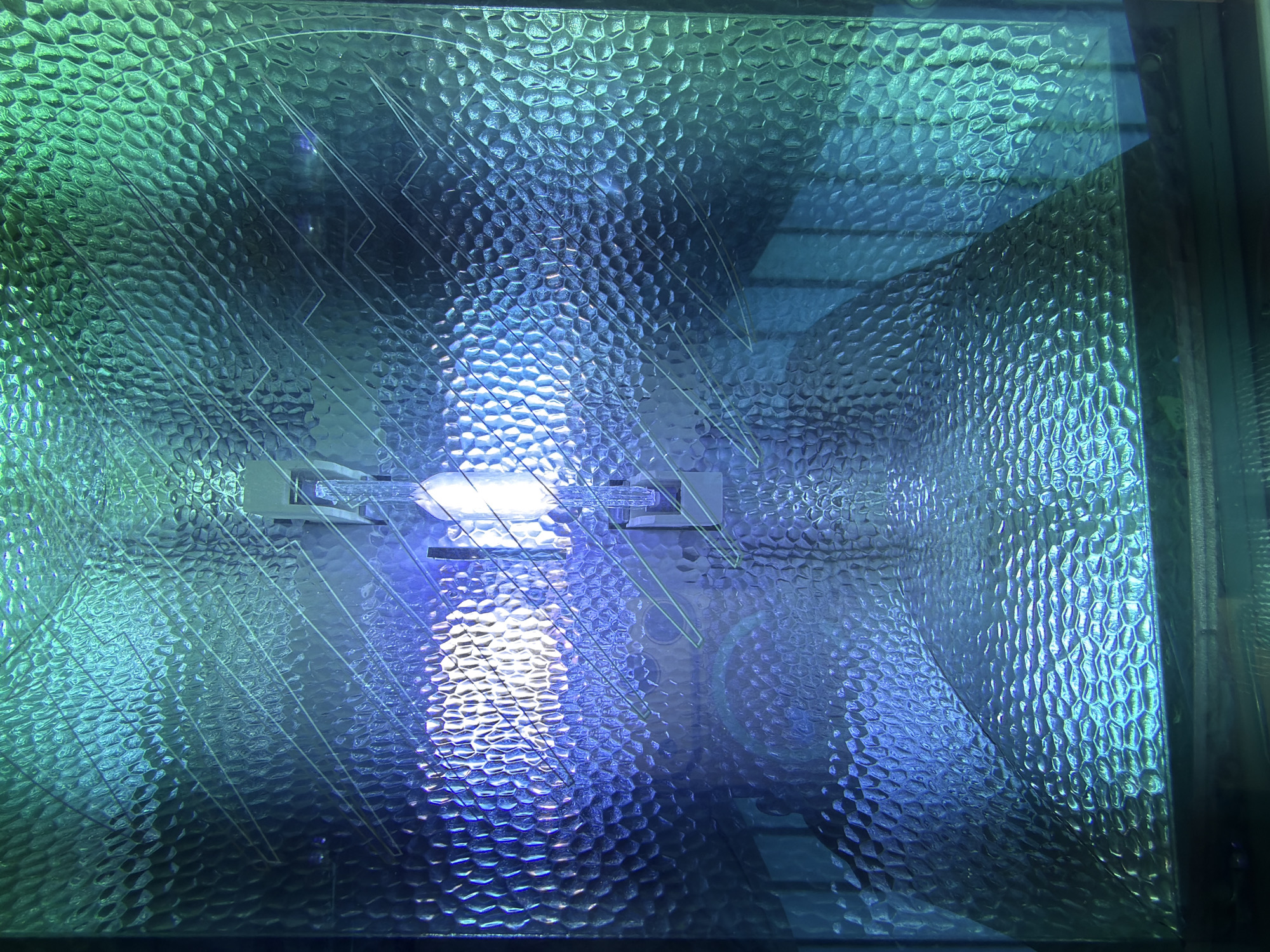 This is a close-up, abstract photograph of the interior of a light fixture, likely a grow light or industrial lamp, seen through a textured glass or plastic cover. The cover has a honeycomb-like pattern that distorts the light source within. The light itself appears to be a bright white or bluish-white, with strong reflections and refractions creating patterns of blue and green light across the textured surface. Faint, thin lines, possibly indicating a circuit board or etched design, are visible on the surface, overlaid by the light patterns. The overall impression is one of intense, artificial illumination with a textured, fragmented visual field. There are no people or any identifiable urban elements visible. The location in Kyiv, Ukraine, cannot be determined from the visual information presented in this image.