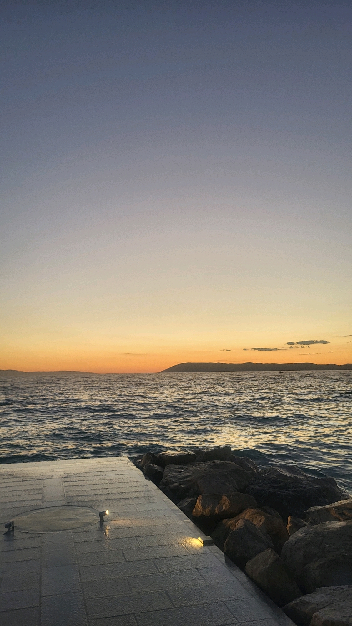 This is a vertical, full shot of a waterfront scene at dusk in Općina Tučepi, Croatia. The sky transitions from a pale blue at the top to a warm orange glow near the horizon. A dark, silhouetted landmass is visible in the distance. The sea is calm with gentle ripples reflecting the twilight colors. In the foreground, there is a paved walkway made of grey rectangular tiles that appears to be wet, with two small, illuminated lights embedded in it. To the right of the walkway, a rocky embankment juts into the sea, composed of large, dark grey stones.