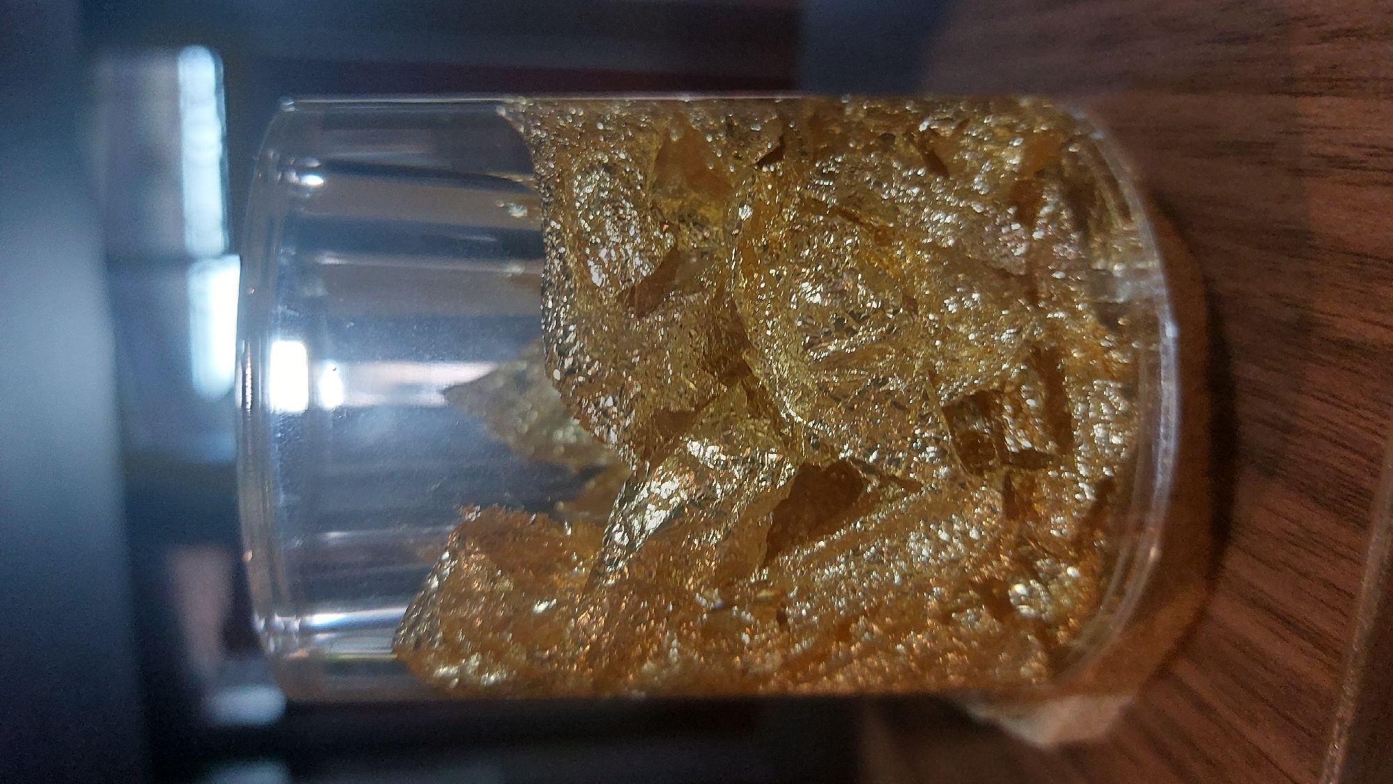 A clear glass container is filled with crumpled gold leaf, which reflects light and creates a shimmering effect. The container is placed on a wooden surface, possibly a table or shelf. The background is blurred and dark, with some out-of-focus lights. The image appears to be a close-up shot of the glass container. The overall impression is one of luxury or opulence due to the use of gold leaf. No specific location in the City of Medan, Indonesia, can be determined from the image.