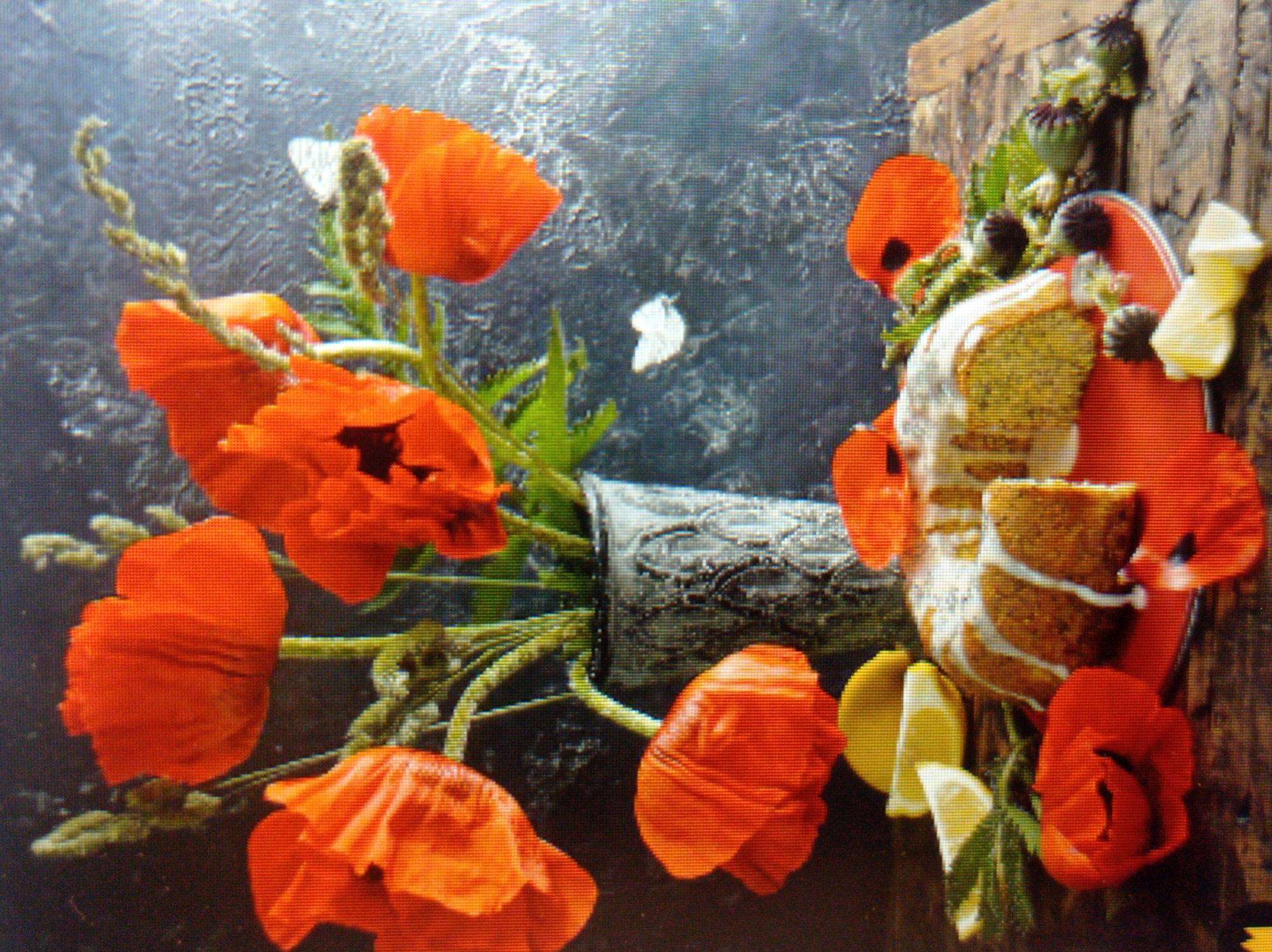 The image depicts a still life arrangement on a wooden surface, featuring bright orange poppies in a decorative vase, a sliced cake drizzled with white icing, lemon wedges, and other botanical elements. A small white butterfly is visible near the flowers. The background is a textured, dark surface. The scene is a photographic still life and does not depict any location in the city of Mukachevo, Ukraine, nor does it contain any people or discernible actions or interactions.