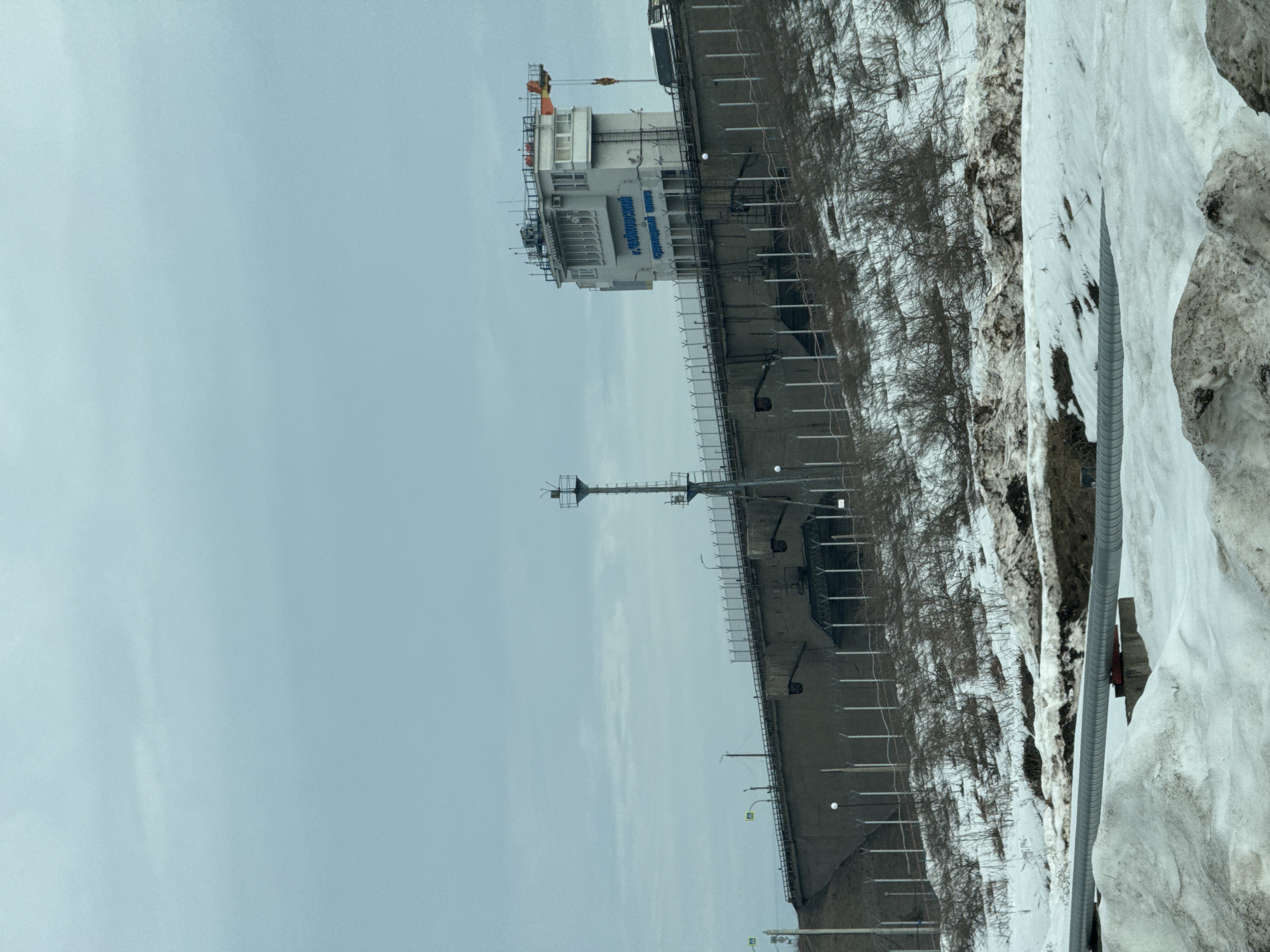 The image depicts a view of the Votkinsk Hydroelectric Station located in the city of Chaikovsky, Perm Krai, Russia. The hydroelectric dam's control building is visible, as well as a portion of the dam structure. The surrounding terrain shows a mix of vegetation and snow cover, likely indicating a winter or early spring season. The sky is overcast. A metallic pipe is visible on the right side of the image.