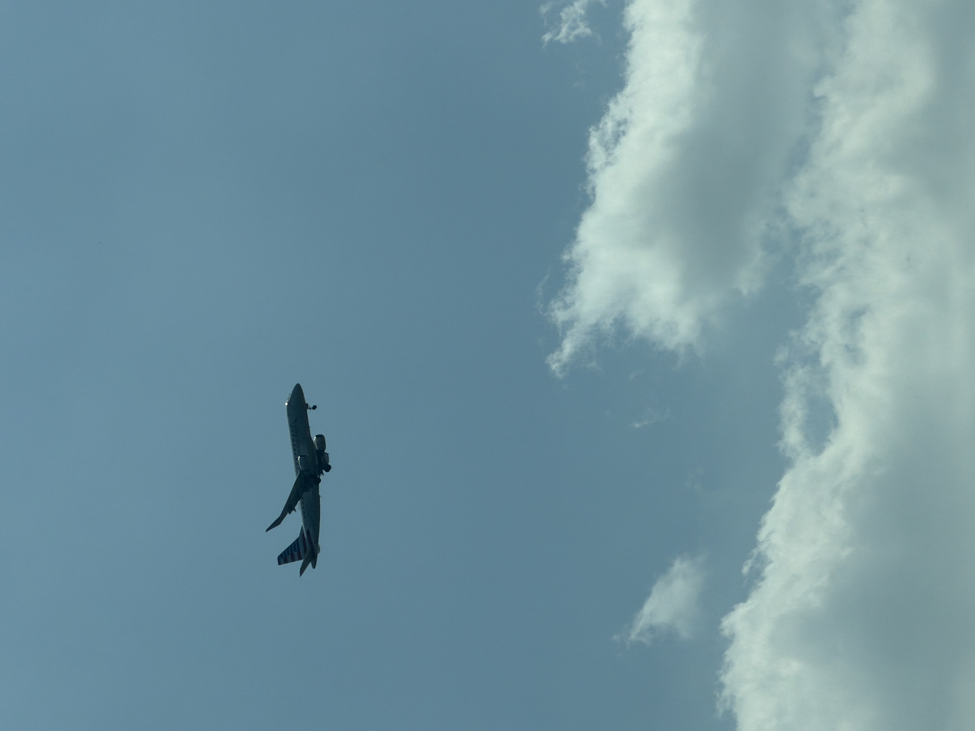 A commercial airplane, identified by the American Eagle logo on its tail, is captured in flight against a clear, pale blue sky. The aircraft appears to be in the process of landing or taking off, with its landing gear visible and extended. Below the airplane, fluffy white clouds are scattered across the lower portion of the frame. The image does not contain any specific landmarks or contextual clues to pinpoint its location within National Harbor, United States of America, nor does it depict any people or interactions.
