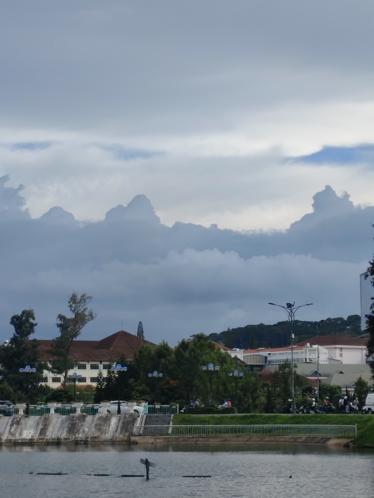 The image captures a cloudy day in Da Lat, Vietnam, near Xuan Huong Lake. In the foreground, the calm, dark water of the lake reflects the overcast sky and the structures along the bank. A portion of a concrete embankment with decorative planters is visible, leading to a pathway lined with trees and streetlights. Beyond the embankment, buildings with terracotta roofs and light-colored facades are nestled amongst dense green trees. A hill covered in dark green trees rises in the background, topped by more buildings and the dramatic, cloud-filled sky. A small group of people on motorcycles are visible on the road behind the embankment, suggesting a typical urban scene. The overall impression is one of a serene, slightly somber atmosphere, characteristic of a cloudy day in this highland city.