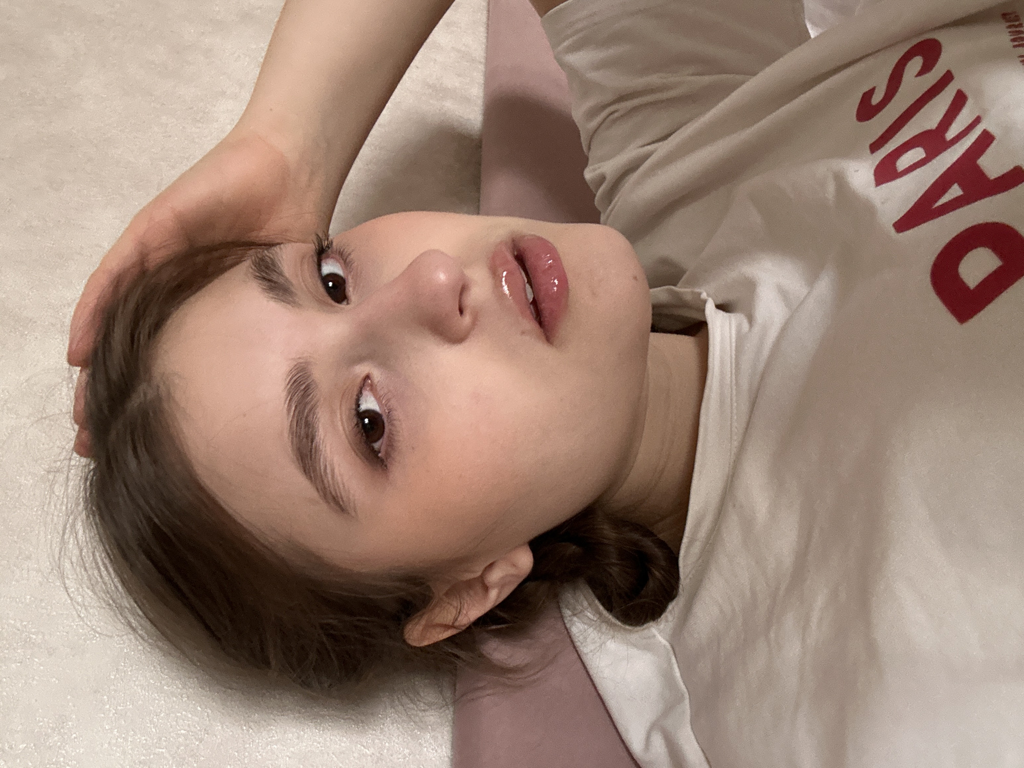 A young female, appearing to be an adolescent or young adult, is depicted in a close-up, chest-up orientation, in a reclining position. Her head rests against a pink fabric surface, with a textured, light-toned wall visible behind her. Her left hand rests on the top of her head, drawing back strands of her brown hair. Her dark brown eyes are open and directed forward. Her lips are slightly parted, revealing a shiny, possibly glossed, surface. She wears a light-toned shirt, with the red capitalized text "PARIS" partially visible on the lower right portion of the garment. The environment is illuminated by ambient indoor light. This image captures a scene within the city of Sarapul, Russia.