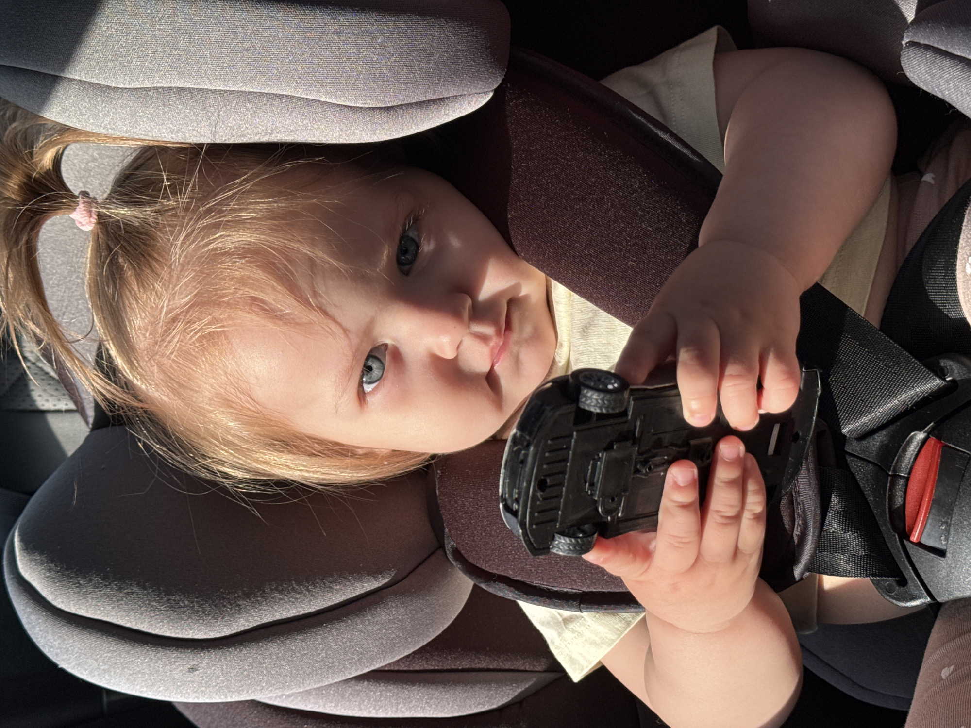A baby with blonde hair pulled back into a ponytail is sitting in a car seat, holding a black toy car upside down in their hands. The car seat is gray and has a black safety harness. The baby has blue eyes and is looking at the camera. The location is not discernible and therefore cannot be determined to be Chaikovsky, Russia.