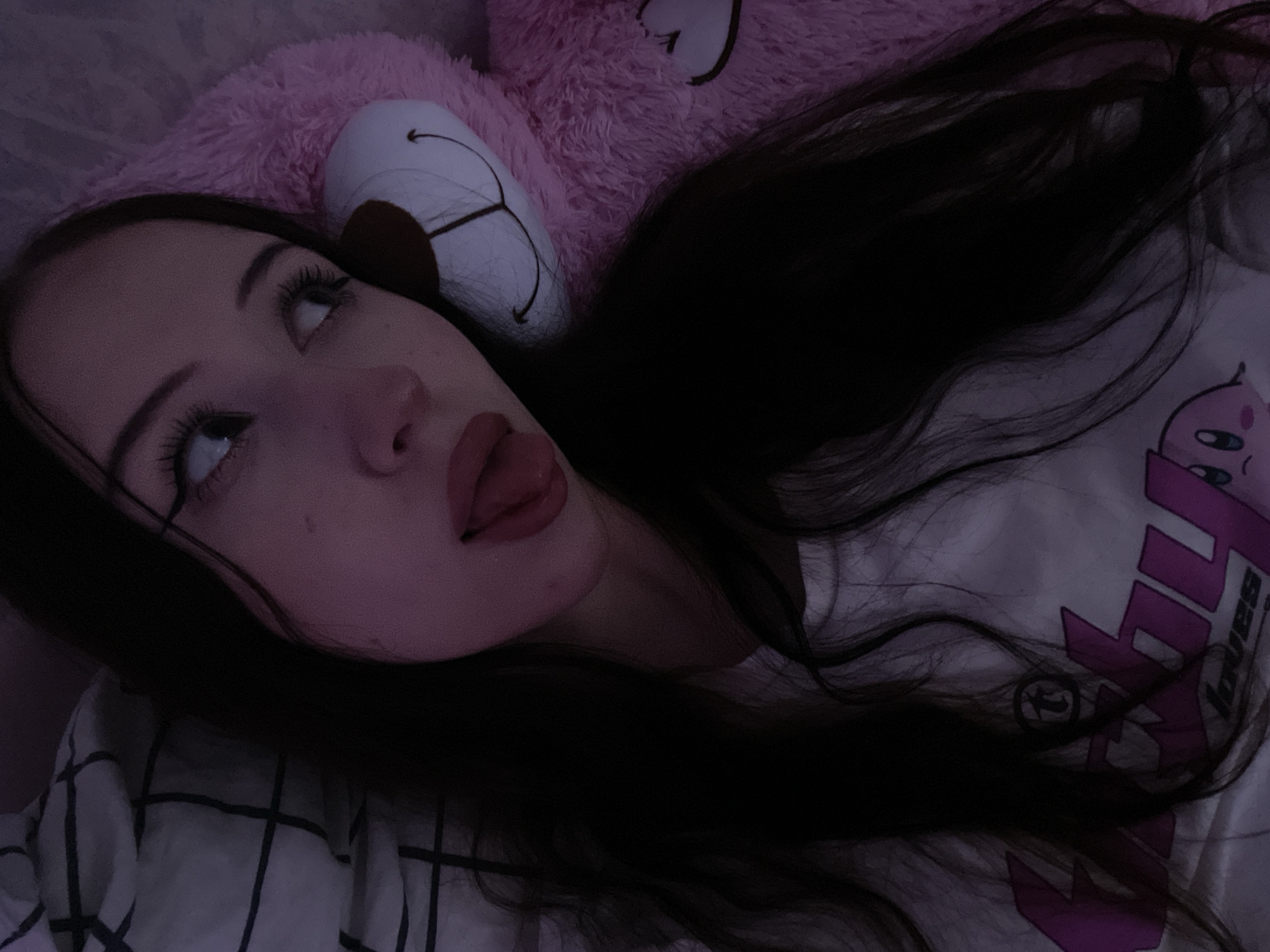 The image shows a young woman lying down next to a large pink teddy bear. The woman has long, dark hair and is wearing a white t-shirt with a graphic print. She is looking to the side with her tongue sticking out slightly. The teddy bear has a friendly expression with stitched eyes and a smile. The background appears to be a bed with checkered bedding. The lighting is dim, with a purple hue, suggesting it might be nighttime or a dimly lit room. There is no discernible information about the location in Votkinsk, Russia.