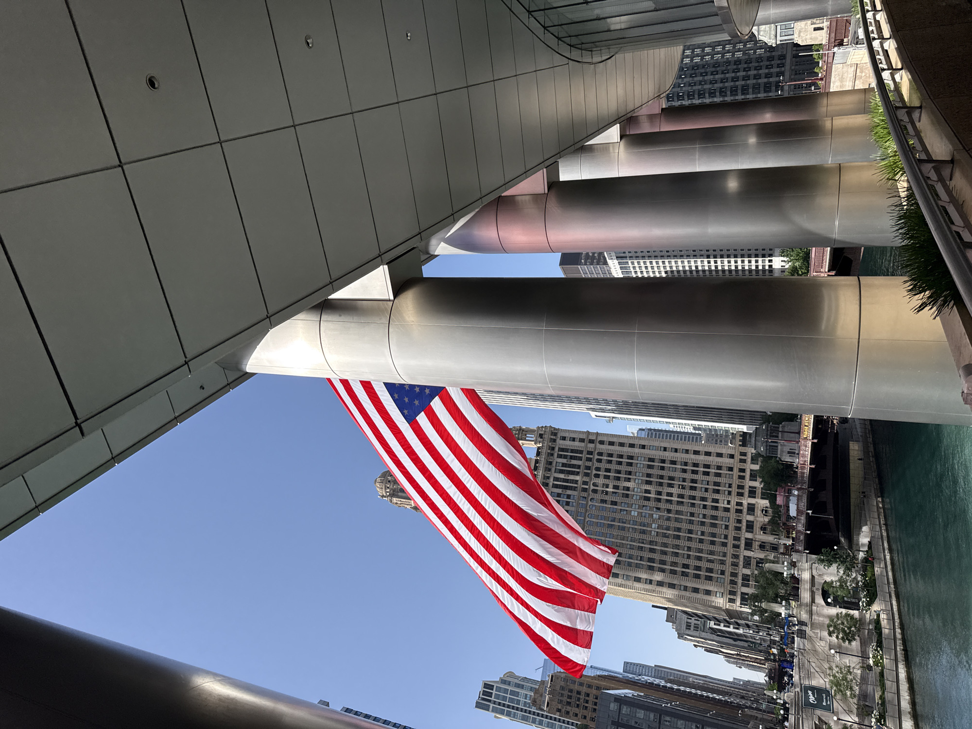 The image captures a view of the Chicago River in the United States of America, likely during daylight hours given the bright sky and clear visibility. The scene is characterized by modern architecture, with a focus on a large, flowing American flag prominently displayed against a clear blue sky. The flag is angled downwards and to the right, its red and white stripes and blue union with stars clearly visible.
The foreground features the curved, metallic underside of a building, with large, cylindrical support structures extending outwards. These structures are highly reflective, mirroring the sky and the surrounding environment. Beyond the flag and architectural elements, a cityscape unfolds, with numerous tall buildings of varying architectural styles and colors forming the urban backdrop. The Chicago River is visible on the right side of the image, its green waters reflecting the urban surroundings.
The overall atmosphere conveyed by the image is one of civic pride and urban dynamism. The bright sunlight and clear sky suggest a pleasant day, ideal for outdoor activities or exploring the city. While no specific event is explicitly depicted, the presence of the flag and the bustling urban environment hint at a lively atmosphere. There is no visible text in the image.