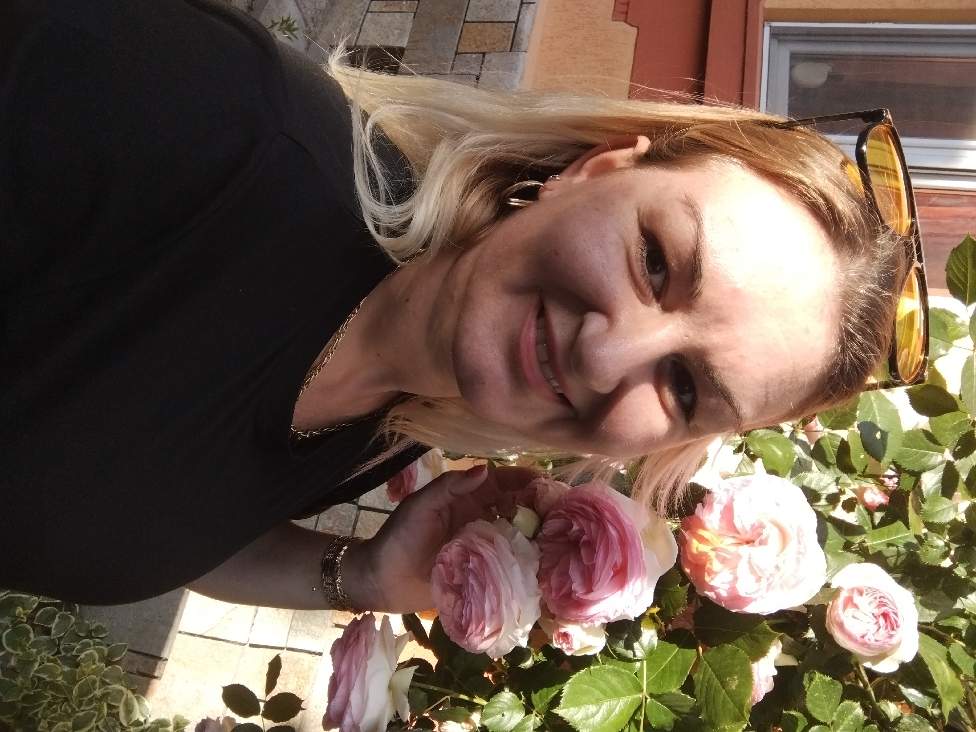 A woman is shown in a close-up shot, standing in what appears to be a garden. She is holding a cluster of light pink and white roses in her left hand. The roses are in full bloom with visible green foliage. She is wearing a black top, gold hoop earrings, and a gold bracelet. A gold chain is visible around her neck. Sunglasses are perched on top of her head. The background includes a brick or stone surface and a building with a white-framed window. The specific location in Stara Zagora, Bulgaria cannot be identified from the image.