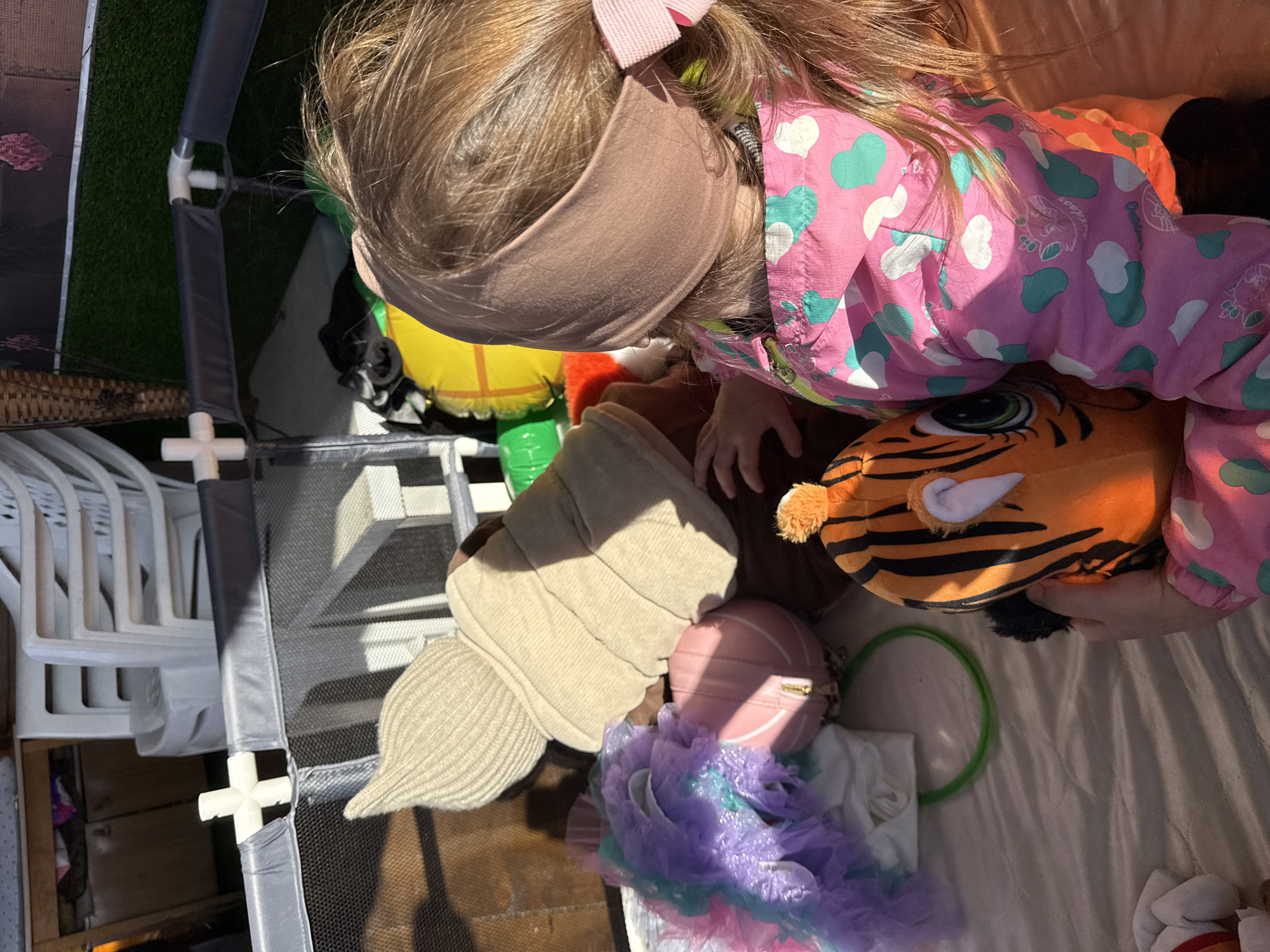 A young child with blonde hair and a pink bow stands indoors, holding a tiger-striped stuffed animal. The child wears a pink jacket with a green and white heart pattern and a brown headband. A tan-colored beanie, a pink purse, and a blue and purple tutu are present on the floor. The indoor location includes a playpen in the top left of the frame, and white plastic chairs are visible nearby. The location is presumably in Кипчак, Russia.
