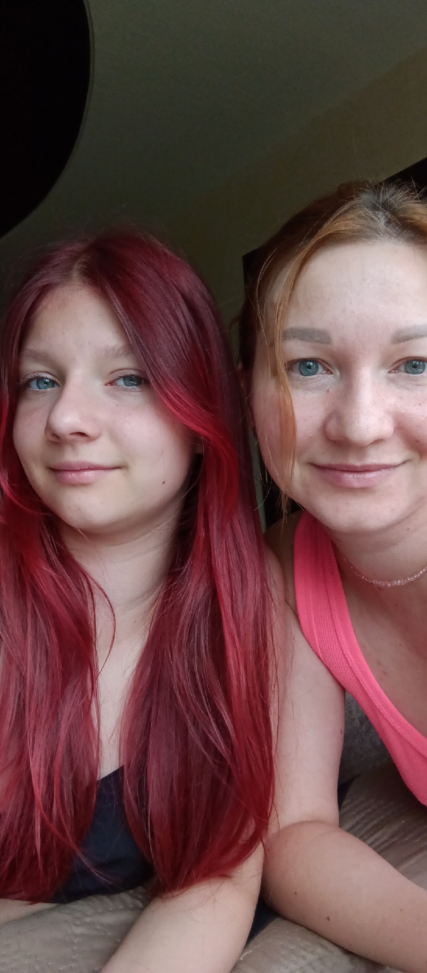 A close-up shot shows two fair-skinned women in the city of Unknown, Unknown. The woman on the left has long, straight, red hair, blue eyes, and freckles on her nose and cheeks. She is wearing a dark-colored top. To her right is another woman with light-brown hair, also with blue eyes and freckles. She is wearing a pink tank top and a beaded necklace. Both women are looking directly at the camera with slight smiles. The background is dark and out of focus.