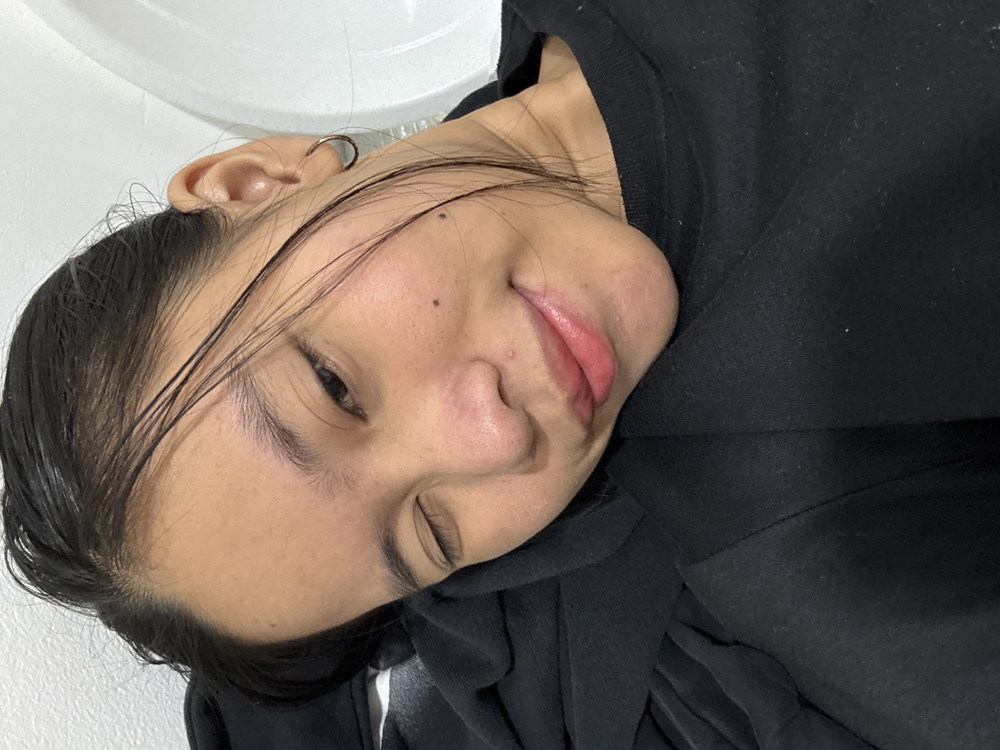 The image is a close-up, selfie-style shot of a young person with dark hair, looking towards the camera. They are wearing a black t-shirt. One eye is closed in a wink, while the other is partially open. Their head is resting on a black fabric, possibly a blanket or clothing. The background appears to be a white wall and a portion of a white sink or bathtub. There are no discernible landmarks or contextual clues within the image to suggest a specific location in South Jakarta, Indonesia. The lighting is soft and appears to be natural or from an overhead source.