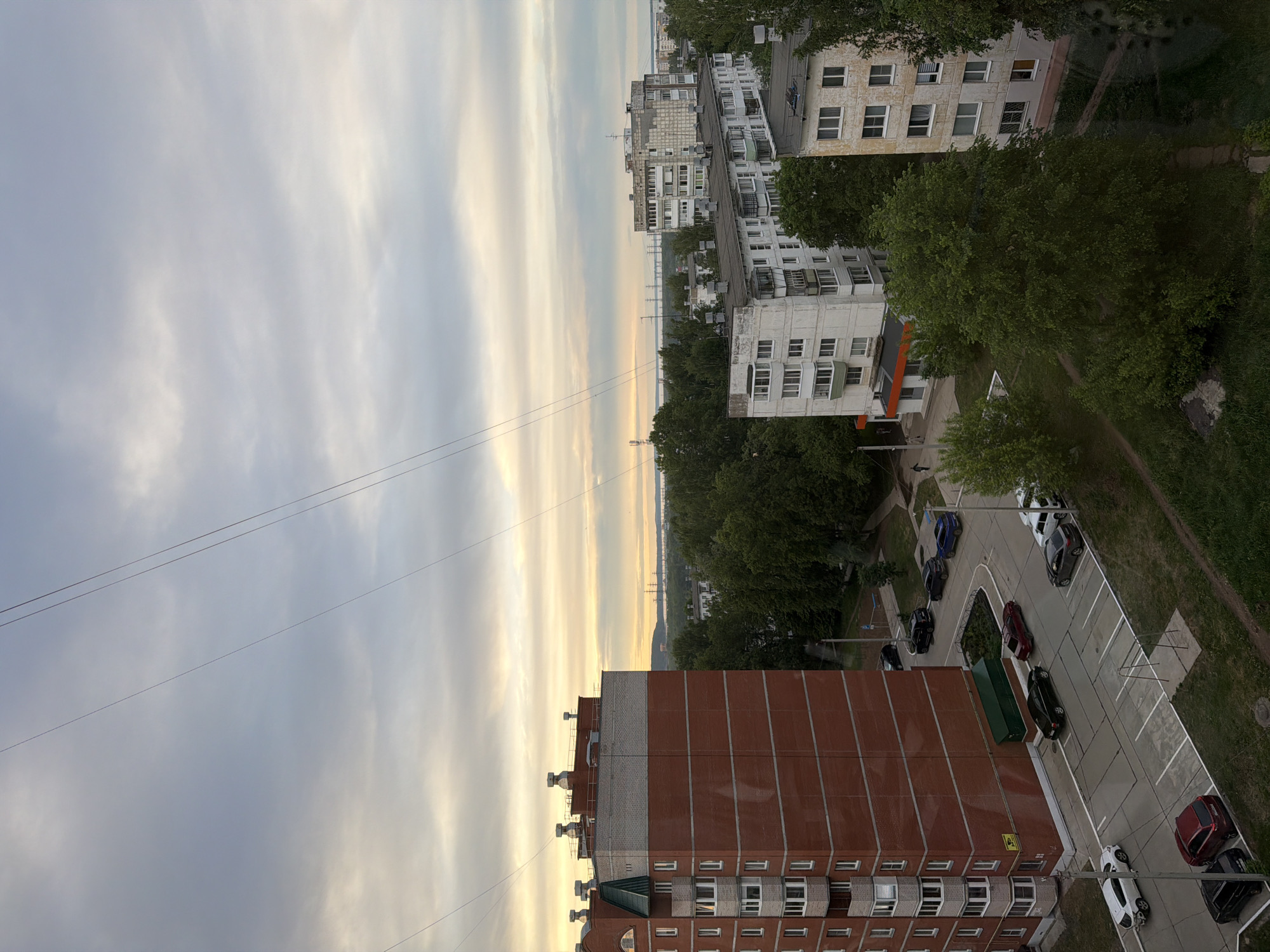 The image shows an aerial view of a residential area in Chaikovsky, Russia. The perspective is from a high-rise building, looking down on a parking lot with several cars, including a red car, a white car, and several black cars. A large brick building with white window frames and multiple floors is prominently featured in the foreground. In the background, there are other apartment buildings of varying heights and designs, interspersed with green trees. The sky is overcast with striated clouds, suggesting either dawn or dusk. Electrical wires are visible in the upper left part of the frame. The presence of a wide body of water, possibly a river or lake, is discernible in the distance.