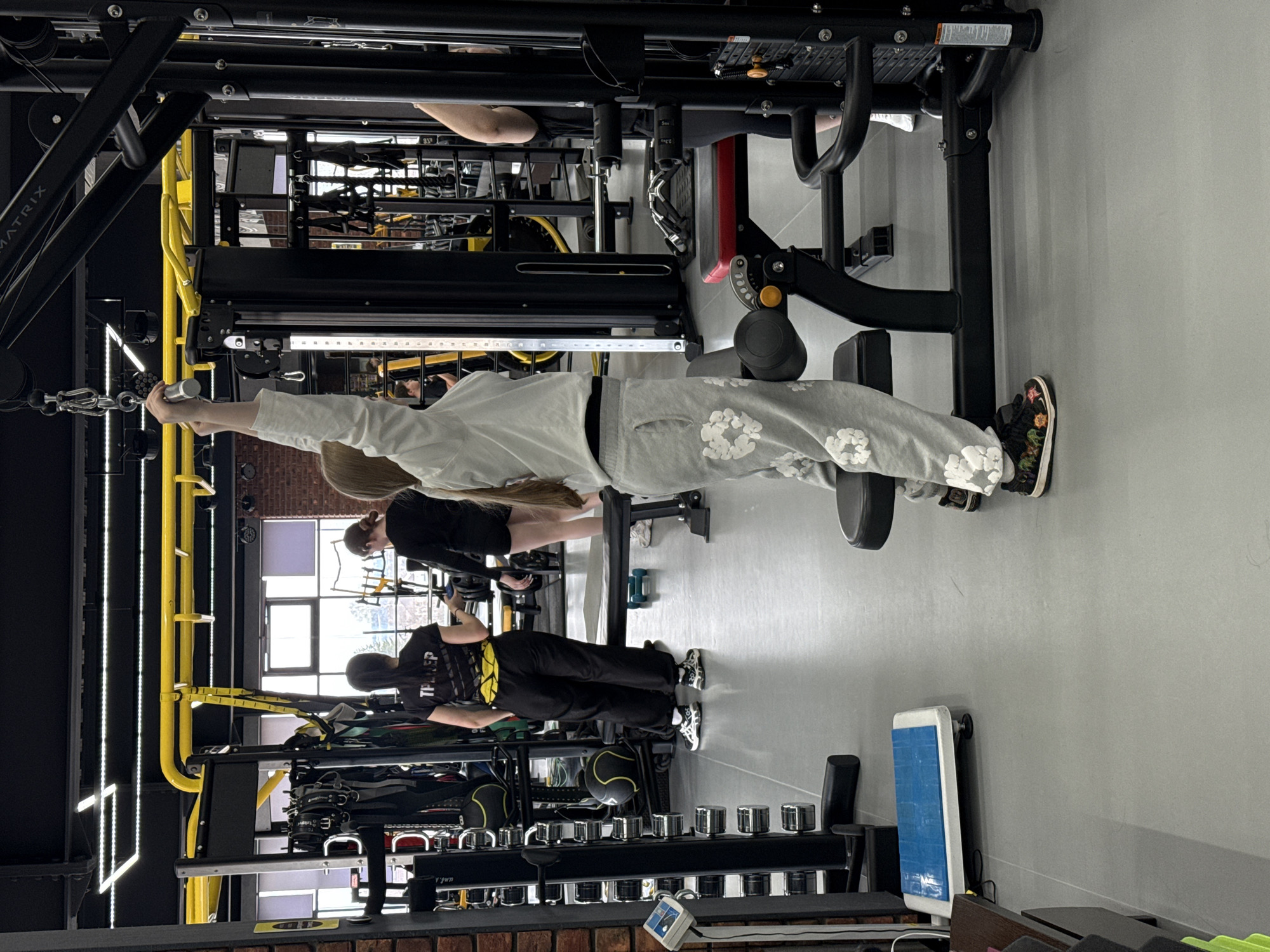 A gym interior is shown with three individuals engaging in various exercises. One person, dressed in a gray sweatsuit adorned with white floral patterns, is positioned at a cable machine. They are pulling the cable towards them with their right arm extended. Further back, another individual is seated, potentially using a different machine. At the lower part of the frame, a third person is observed using a separate gym apparatus. The environment is filled with various weightlifting machines and equipment. The location is specified as Votkinsk, Russia.