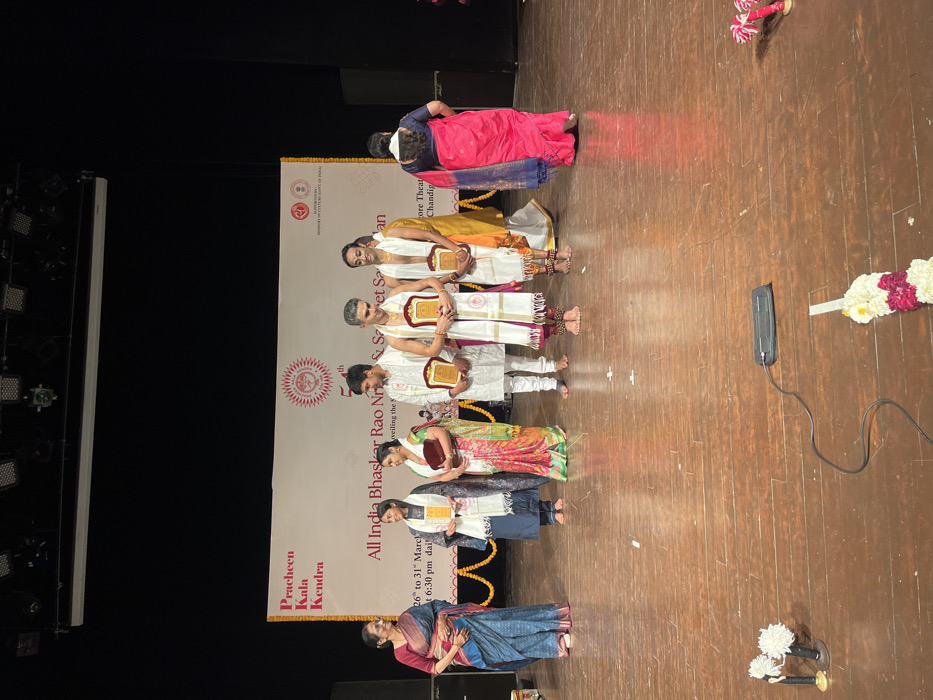 The image shows a group of seven people standing on a stage, facing the audience. They are all dressed in traditional Indian attire. There is a woman in blue and green saree at the left, followed by two women in saree, all holding certificates. Next, there are four men standing in a row in white and yellow dhoti, also holding certificates. The woman at the right is wearing a pink and blue saree. The stage is decorated with garlands of white and pink flowers. The location is the "Pracheen Kala Kendra", which is at the coordinates (latitude: 30.733, longitude: 76.789) in the city of Chandigarh, India. The stage has a wooden floor. The event appears to be an award ceremony for a competition or festival. The banner behind them reads "All India Bhaskar Rao Nritya & Sangeet Sammelan" and "Pracheen Kala Kendra". A black speaker is on the floor in front of the stage. The lighting is bright and the atmosphere is festive. The people are smiling and appear to be happy to be part of the event.