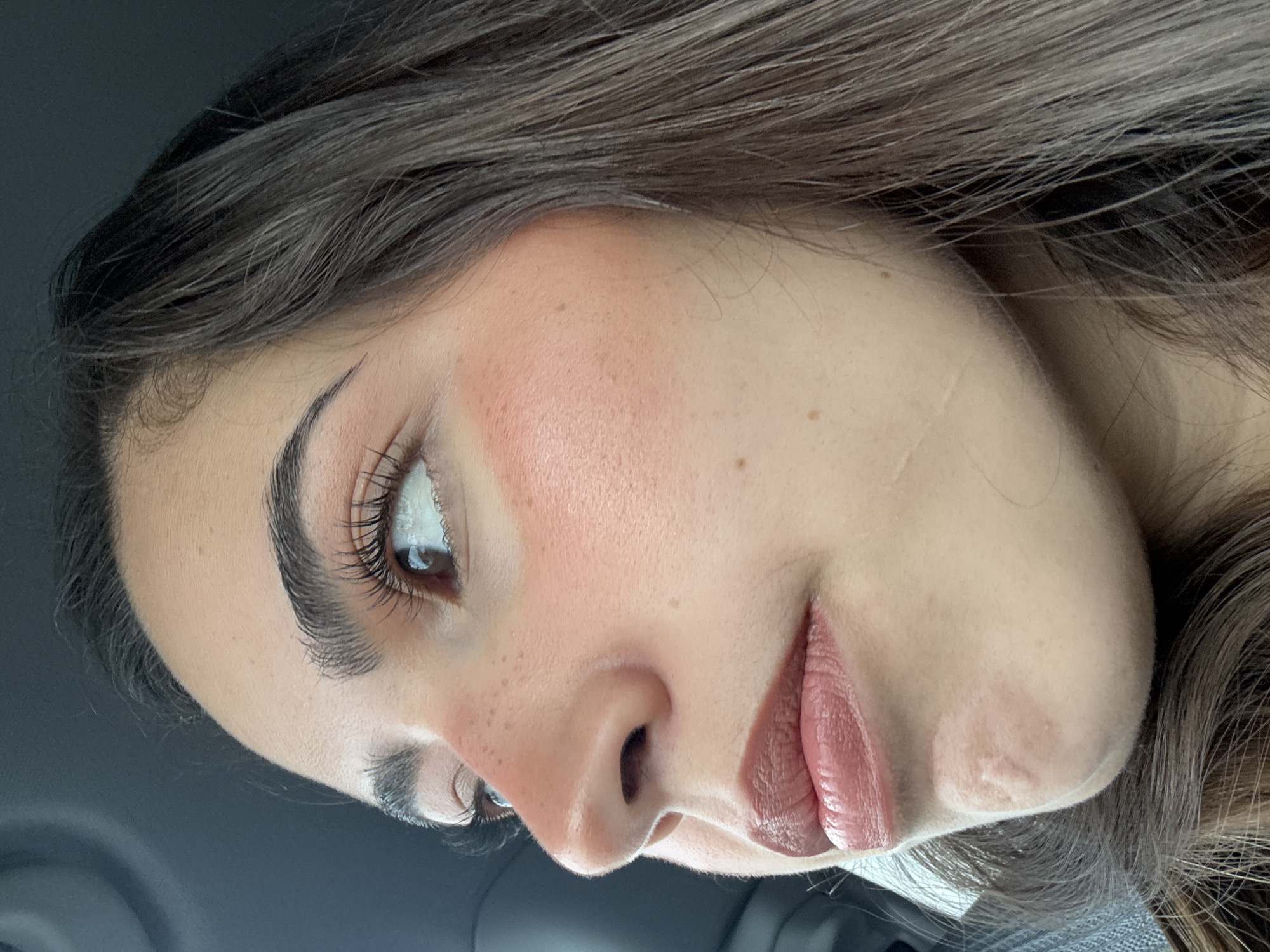 A close-up, rotated portrait of a woman's face, showing her features in detail. Her hair is dark brown with lighter highlights. She has dark eyebrows that are meticulously shaped and long, dark eyelashes. Her skin is fair with rosy cheeks. She has a few freckles on her nose and cheeks. Her lips are painted with a nude shade of lipstick. The background is dark, suggesting she is possibly inside a car. There is no indication from the image that the photo was taken in New Braunfels, United States of America.