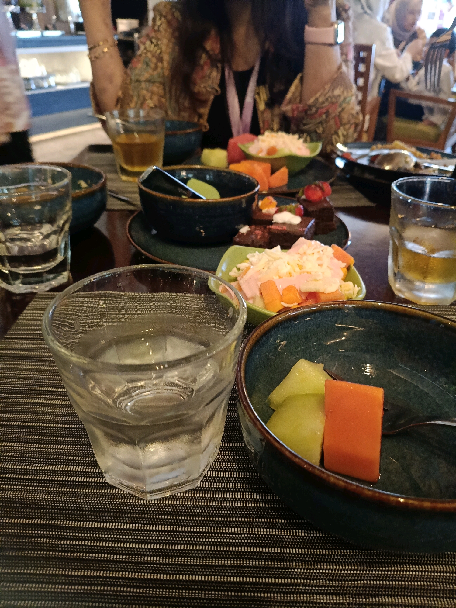 The image captures a close-up view of a table laden with food and drinks, likely at a restaurant in North Jakarta, Indonesia. In the foreground, a glass of water with ice sits on a dark, textured placemat. To its right, a blue bowl contains diced cantaloupe and what appears to be papaya. Behind this, another blue bowl holds a piece of chocolate cake topped with whipped cream and a strawberry, alongside a salad of shredded cheese, diced carrots, and what looks like ham or fruit in a green bowl. A glass of amber liquid, possibly tea or juice, is positioned to the right of the cake and salad. In the background, out of focus, are several people, including a woman in a patterned top wearing bracelets, and others seated around tables, suggesting a dining setting. The overall ambiance appears to be casual dining.