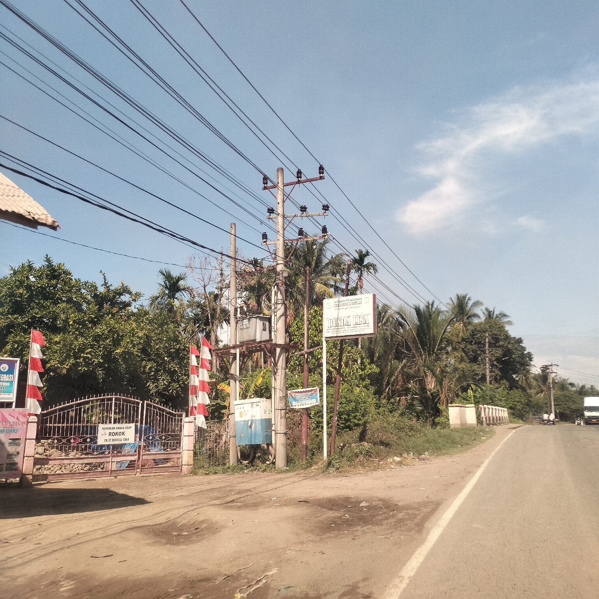 The image captures an outdoor scene in Manetunong, Indonesia, showing a road curving to the right under a clear blue sky with sparse white clouds. The left side of the road features a dirt shoulder, while both sides are lined with abundant green foliage and tropical palm trees.
In the foreground on the left, a metal gate, possibly an entrance to a property, is adorned with numerous red and white flags, characteristic of Indonesia. Partially visible to the left of the gate is a roof structure. A large utility pole stands prominently near the center, equipped with a transformer, insulators, and multiple cross-arms supporting a complex network of power lines that crisscross the upper part of the frame.
Several signs are visible:
- On the utility pole, a white sign reads "YAYASAN PENDIDIKAN BUNDA LINA" (Bunda Lina Education Foundation).
- Below it, a smaller blue sign is present.
- On the gate, a white sign states "KAWASAN BEBAS ASAP ROKOK TK IT BUNDA LINA" (Smoking-free Zone Bunda Lina Kindergarten).
- A partial sign on the far left includes text like "EGRASI NONG ATU RA" and "INDA LINA ID DARU."
The scene is bathed in bright daylight, suggesting a sunny day. In the distance along the road, a white truck and other faint vehicle shapes can be seen. The overall impression is one of a typical rural or semi-urban landscape in a tropical region.