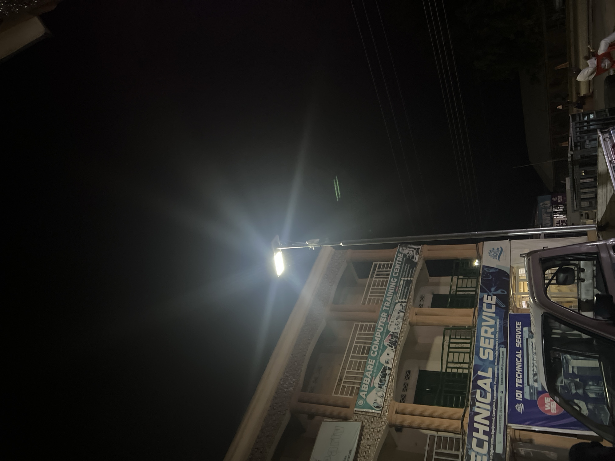 The image depicts a nighttime scene in Jalingo, Nigeria, likely in an urban area. A two-story building dominates the left side of the frame, illuminated by a bright street lamp situated on the right. The building has a cream-colored facade with decorative elements and balconies. Several signs are visible on the building. The top sign reads "ABBARE COMPUTER TRAINING CENTRE" and displays images of computers and students. Below this, a larger blue sign announces "TECHNICAL SERVICE" and "IDI TECHNICAL SERVICE," with contact information and a logo. A portion of a light-colored pickup truck is visible in the foreground, parked in front of the building. Several people are present, including a man sitting on a red chair in the lower right, appearing to be looking at his phone. Utility wires are strung across the dark sky. The overall environment appears to be a commercial street at night, with the buildings and signs indicative of businesses operating in the area.