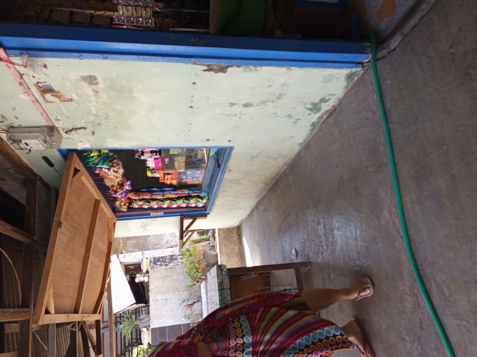 The image shows the exterior of a small shop in Tebel, Indonesia. A person, partially visible on the left, is wearing a colorful patterned dress and flip-flops. The shop has a window display filled with various items, and a wooden awning is propped open above it. The building's walls are weathered, with peeling paint. A green hose lies on the ground in front of the shop. In the background, there are potted plants and other structures, suggesting a semi-urban or rural setting. The overall impression is one of a simple, functional storefront operating in a warm climate.