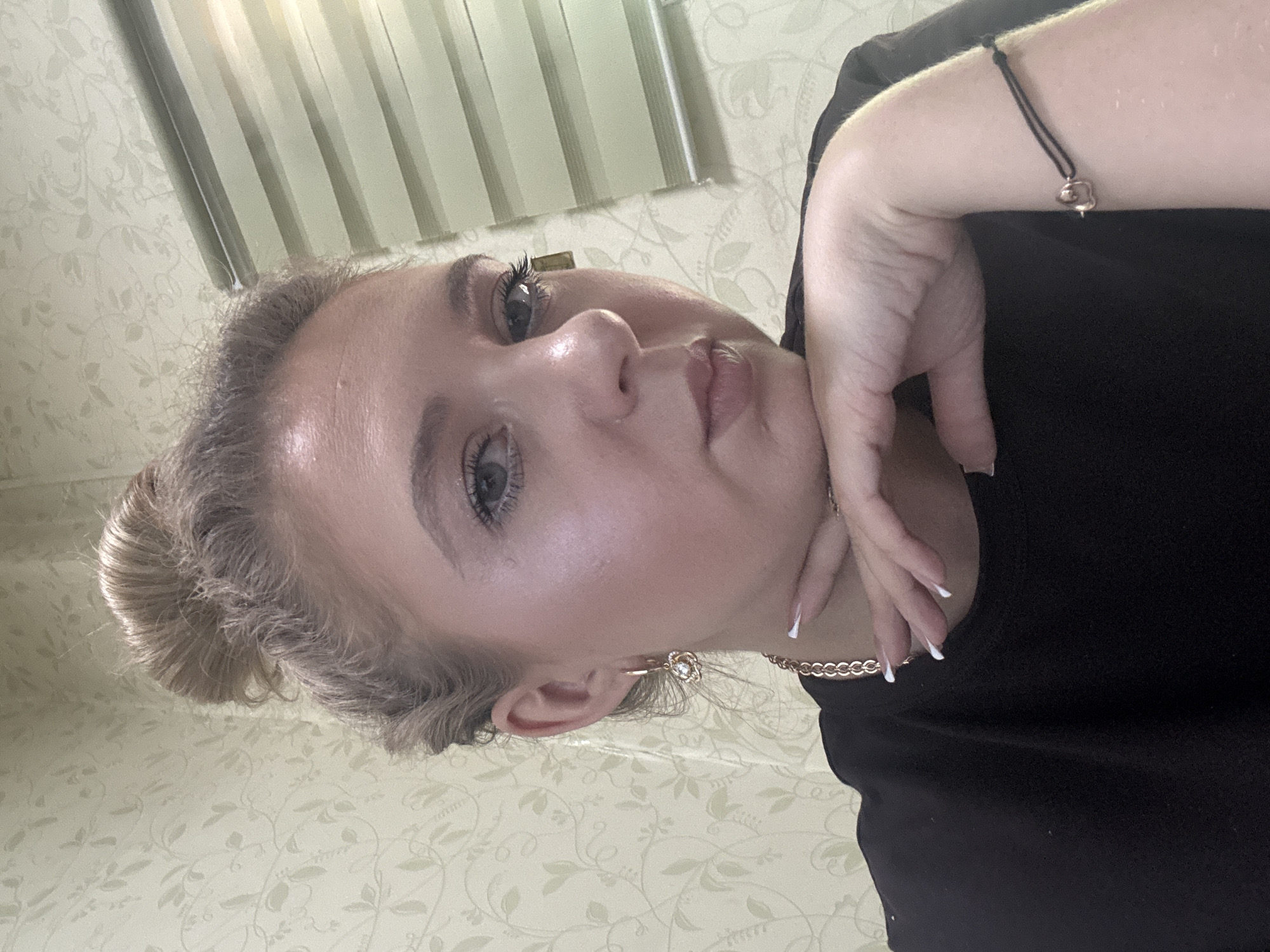 A woman with blonde hair styled in a messy bun rests her chin on her hand. She is wearing a black t-shirt, a rose gold chain necklace with a heart charm, and a rose gold earring with a flower design. Her nails are painted white. The background features a textured wallpaper with a floral pattern and a window with horizontal blinds. The lighting suggests an indoor environment.