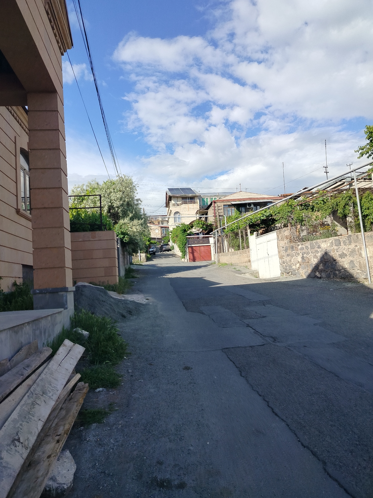 The image depicts a quiet residential street in Yerevan, Armenia. The street is paved with asphalt, showing signs of wear and patching. Buildings line both sides of the street, varying in height and architectural style. One building on the left exhibits a light brown stone facade with layered decorative elements. A pile of gravel and some discarded planks lie adjacent to the building. The building on the right has a stone wall covered with vines and a metal framework. In the distance, more houses are visible, some with solar panels on their roofs. Parked cars are seen further down the street. Overhead electrical wires cross the sky, and the sky is partly cloudy with blue patches.