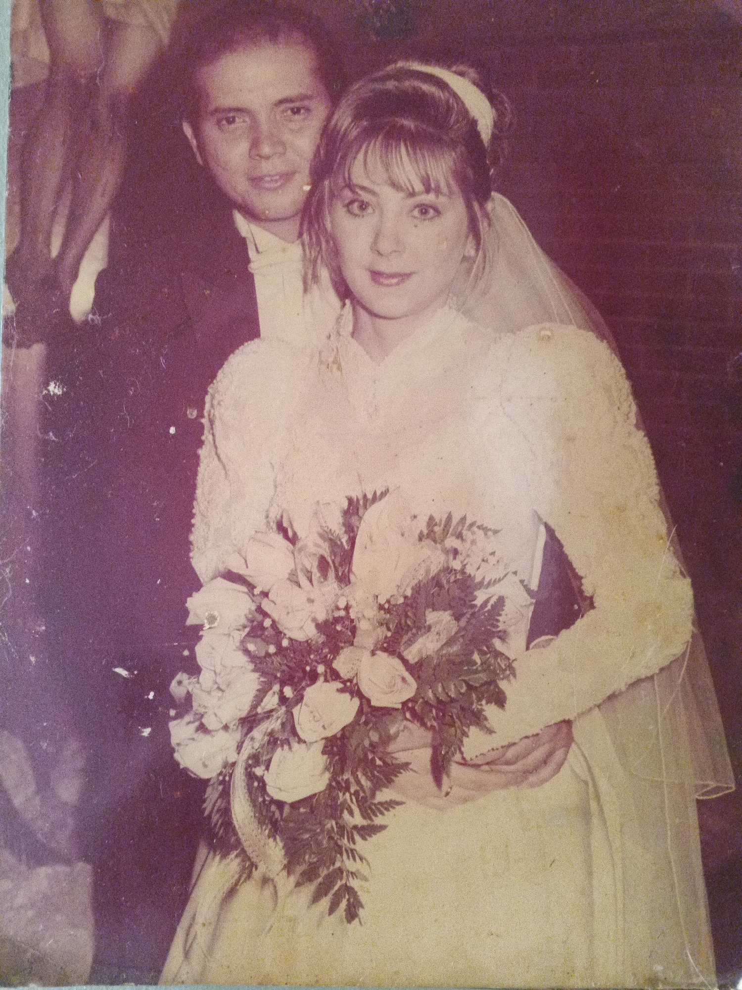 A faded, slightly damaged photograph depicts a wedding scene. A bride, with fair skin and light hair styled with bangs and a headband, is centrally positioned. She wears a long-sleeved wedding gown and a veil, and holds a bouquet of light-colored roses and greenery. A groom stands behind her, partially obscured, wearing a dark suit and light shirt. The location, Caracas, Venezuela, is inferred by the background. The photograph's aged condition suggests a historical image.