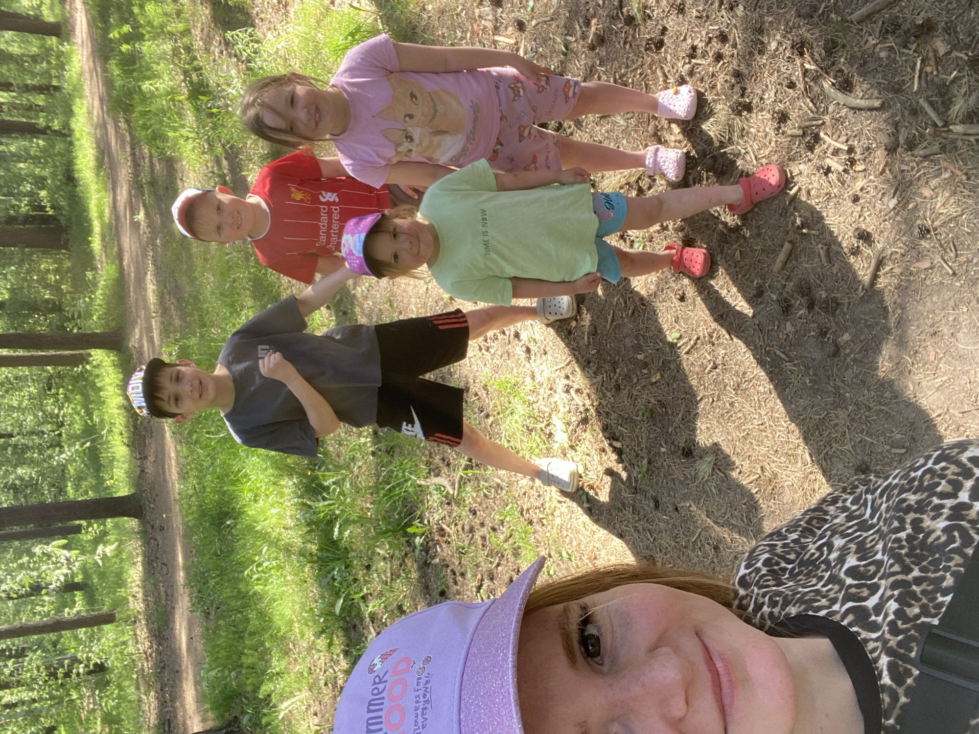 The image shows a selfie of a fair-skinned adult woman with auburn hair, wearing a glittery purple baseball cap and a leopard-print shirt. She is positioned in the foreground. Behind her are four children, presumably her family, standing in a line. The children appear to be of varying ages. The first child wears a red soccer jersey, the second wears a light green T-shirt and a lilac hat, the third wears a cat print pajama and the fourth wears a gray T-shirt. They are standing on a dirt path surrounded by grass and trees. The lighting suggests it is daytime. There is no evidence to indicate that the picture was taken in Chaikovsky, Russia.
