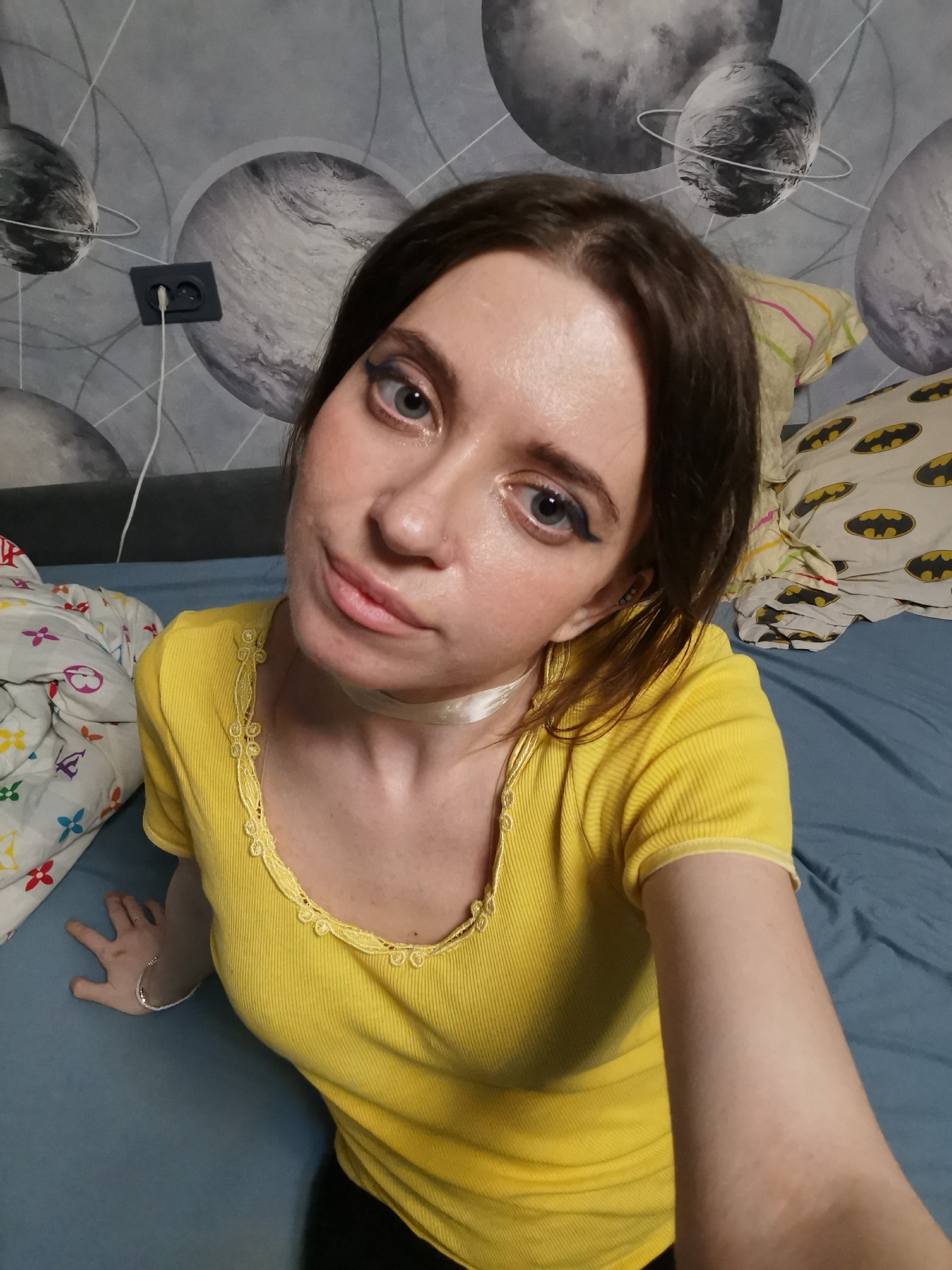 A young woman with brown hair and blue eyes is sitting on a bed. She is wearing a yellow shirt and a white choker. The bed has blue sheets and several pillows with different patterns, including batmans. The wall behind her has a wallpaper with planets. An electrical outlet is visible on the wall. The location appears to be a bedroom. The city is unknown.