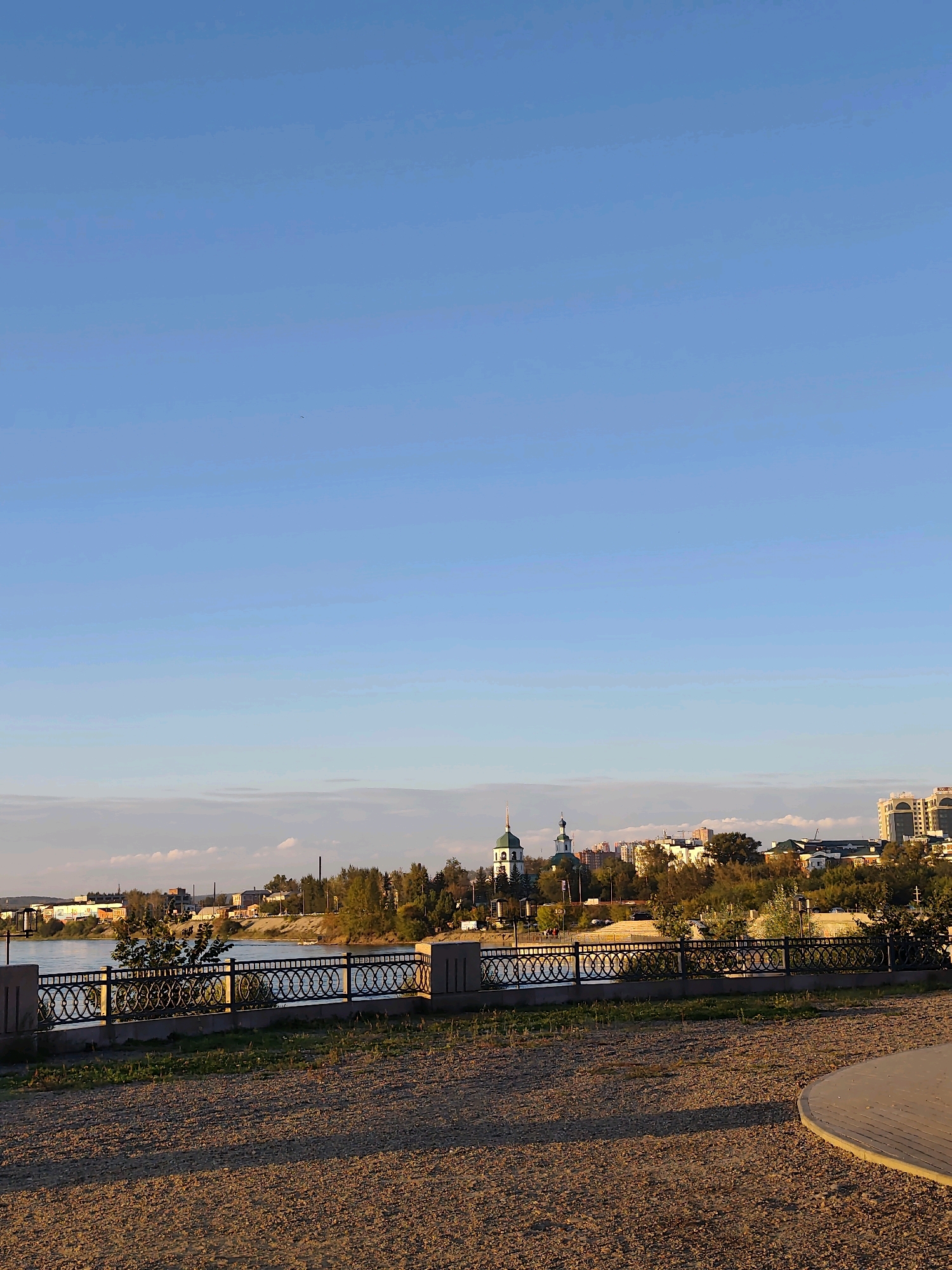 This is an outdoor, horizontal, medium-shot photograph taken from a gravelly area in a city park. In the foreground, the gravelly ground occupies the lower third of the frame, with a curved, light-colored paved area on the right. A black metal fence with an ornate circular pattern runs horizontally across the middle ground, separating the park area from a body of water. Beyond the fence, a river is visible, with a tree growing from its bank.
On the opposite bank of the river, a city skyline is visible. The skyline features various buildings, including a prominent white and green domed church with a spire, a tall modern residential building, and other smaller structures. Green trees are interspersed throughout the cityscape. The sky is a clear, pale blue, with a few thin, wispy clouds near the horizon. The overall lighting suggests it is late afternoon or early evening, with the sun casting long shadows across the gravel. The scene depicts a view of the Angara River in Irkutsk, Russia, with the Holy Trinity Church being a notable landmark in the city's skyline. There are no people visible in the image.