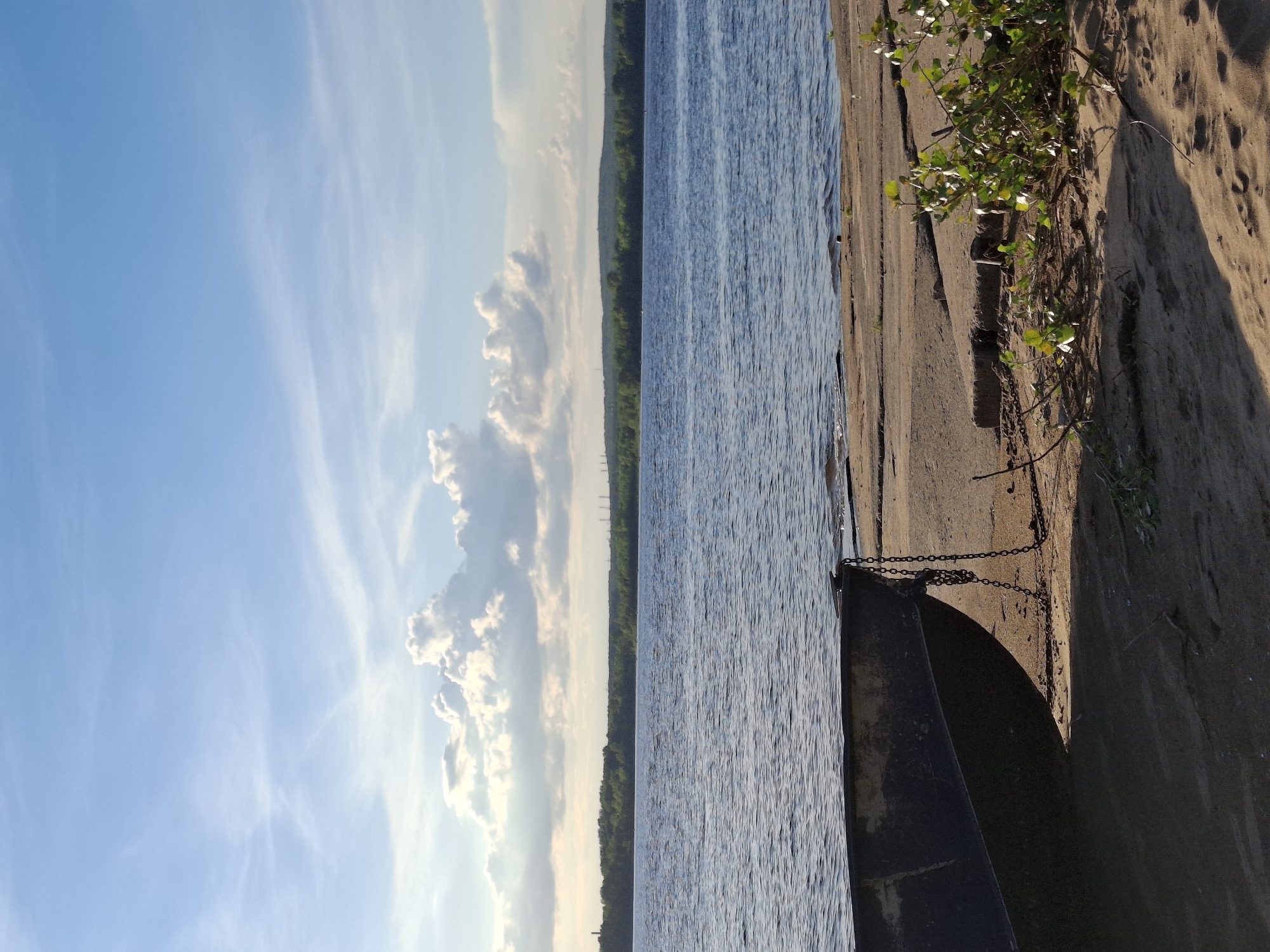 The image depicts a calm river scene in Chaikovsky, Russia, under a bright blue sky with scattered clouds. A dark-colored boat rests on the sandy shore, tethered by a chain. The river stretches across the frame, with a distant tree-lined horizon. The foreground shows sand and some vegetation. The lighting suggests it is either early morning or late afternoon.
