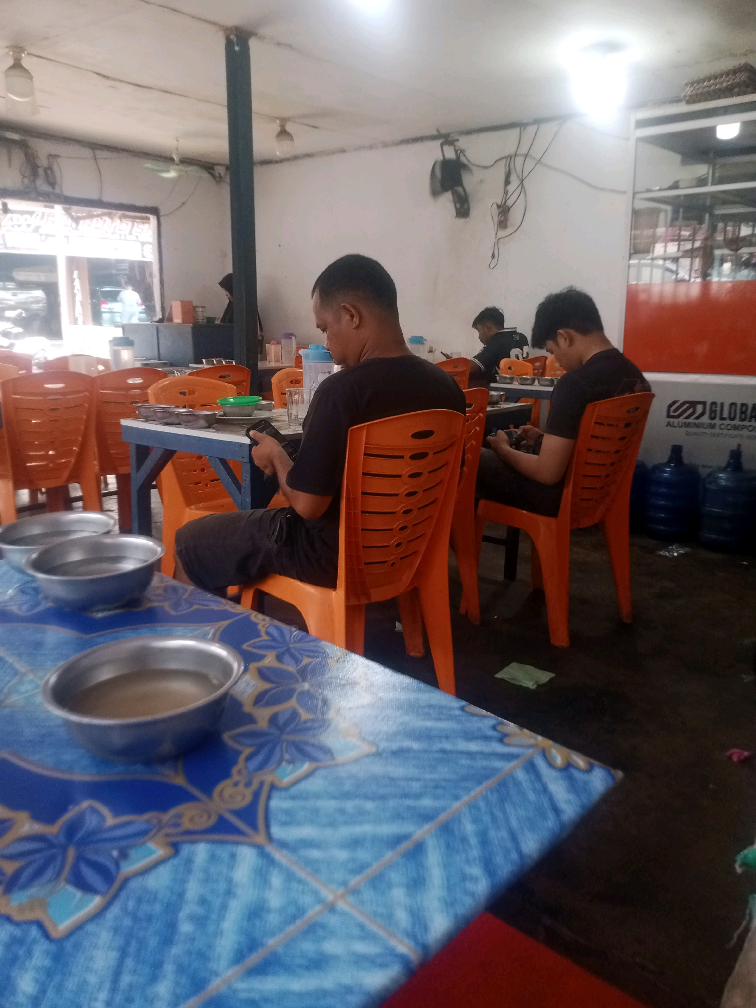 The image shows an indoor scene at a restaurant or eatery. In the foreground, a blue and white patterned tablecloth covers a table, on which several metal bowls containing a light brown liquid are placed. Two people are seated at tables in the middle ground, both facing away from the camera. The person on the left, wearing a black t-shirt, is looking down at a phone. The person on the right, also in a black t-shirt, is similarly engaged with a phone. Both are seated in orange plastic chairs at blue-legged tables. In the background, more orange chairs and tables are visible. The walls are white, and there is a large window or opening with some objects visible behind it. Overhead, fluorescent lights illuminate the space. The flooring appears to be dark and possibly concrete. In the far background to the right, several large blue water bottles and a sign with the word "GLOBAL" are visible, suggesting a commercial establishment. The overall impression is of a casual dining environment. While Pekanbaru, Indonesia is mentioned, there are no specific visual cues in the image to confirm this location.