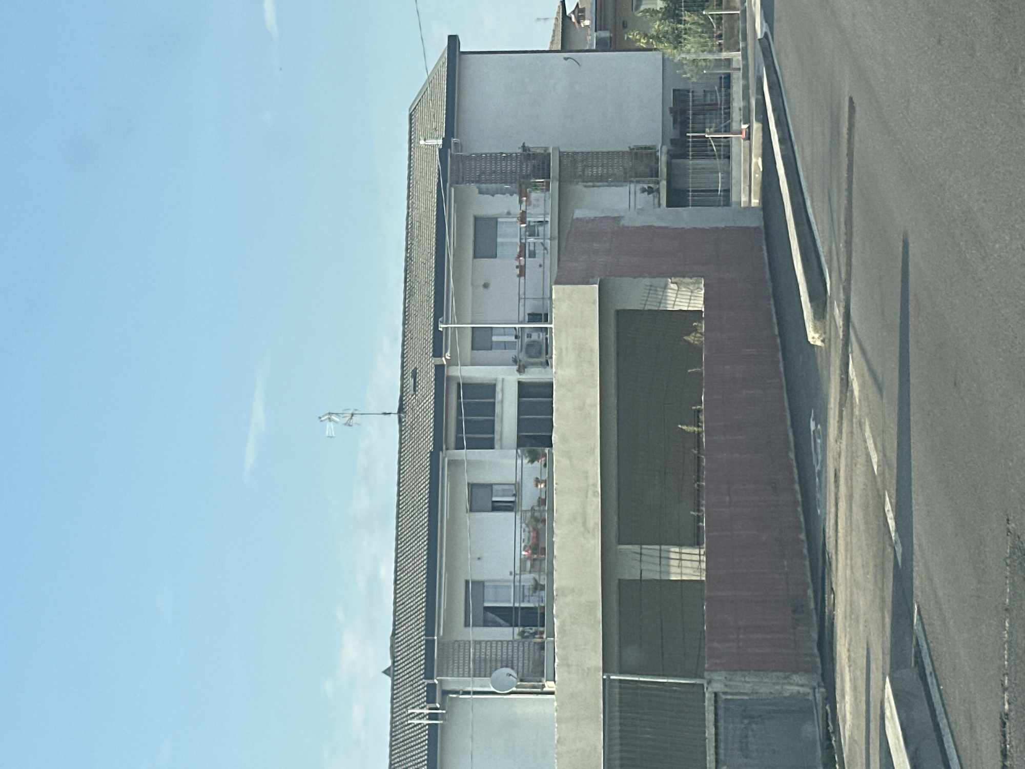 The image shows a view of a residential building with balconies, windows, and a rooftop antenna, situated next to a street. The building appears to be made of concrete and brick. The street is paved and has white markings. The sky is blue with some wispy clouds, suggesting a clear day. The overall scene is urban or suburban, likely residential, given the building's appearance. There are no people visible in the image. The time of day appears to be daytime, given the lighting and shadows. The location is identified as Chiari, Italy. There is no visible text in the image.