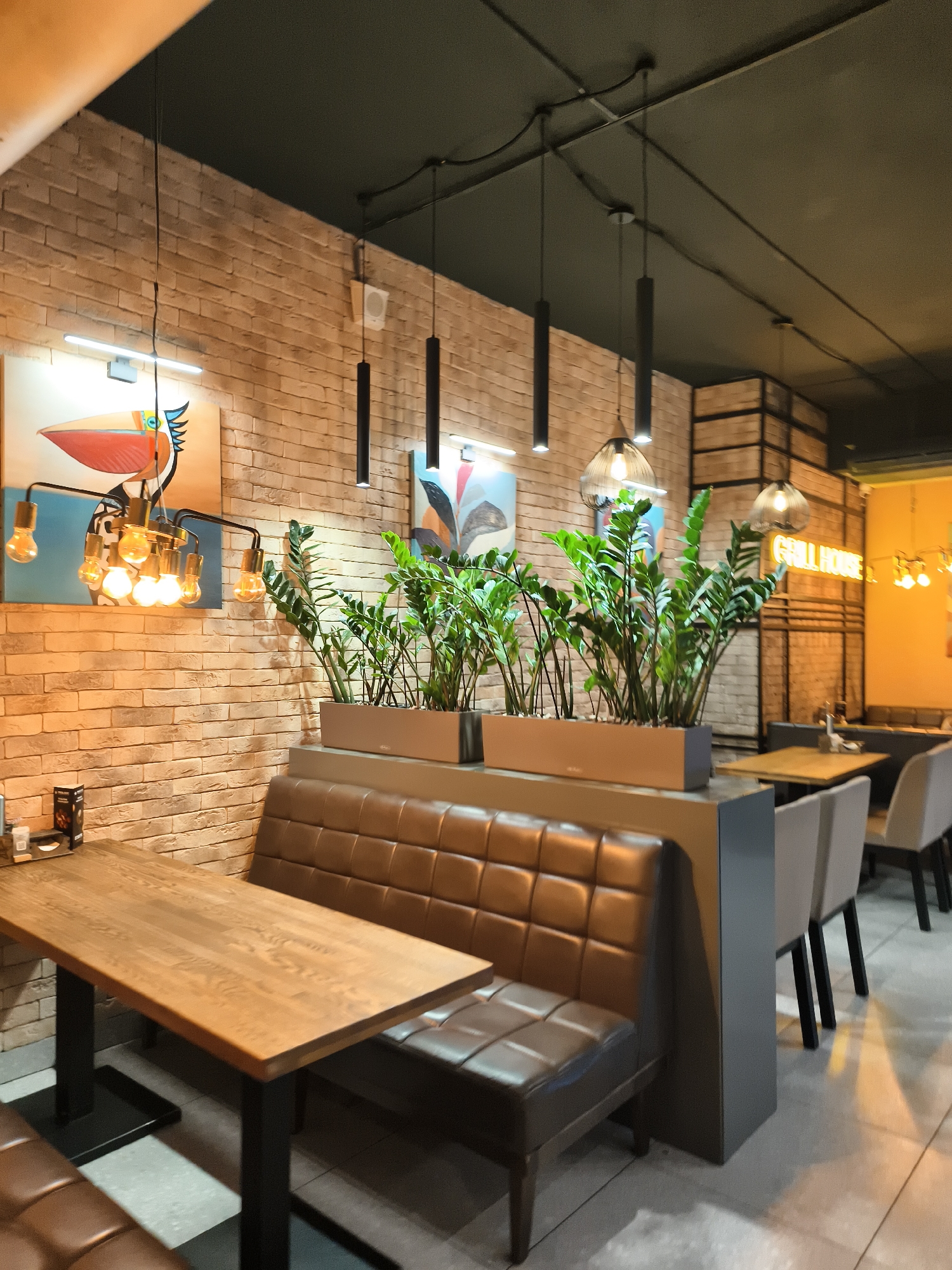 The image depicts an indoor dining area with a rustic yet modern aesthetic. The walls are clad in light-colored brick, contrasting with the dark ceiling and hanging light fixtures. A large wooden table with black legs occupies the foreground, positioned alongside a brown leather booth with tufted seating. Behind the booth, a planter box filled with lush green plants acts as a divider.
Further into the restaurant, more tables and chairs are visible, including light-colored upholstered chairs with black legs. A neon sign that reads "GRILL HOUSE" is prominently displayed on a wall in the background, illuminated by a bright yellow glow. The overall ambiance suggests a casual dining establishment, likely a restaurant or grill. There are no people visible in the image.
